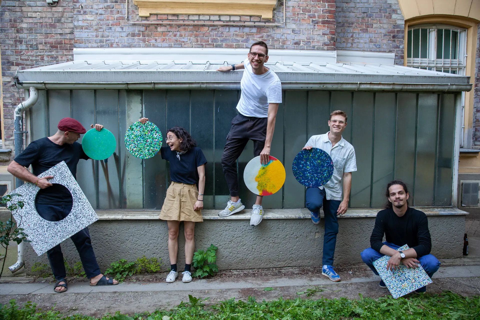 Vier Männer und eine Frau stehen in einem Garten vor einer Wand und halten farbige kreisförmige Paneelen in der Hand