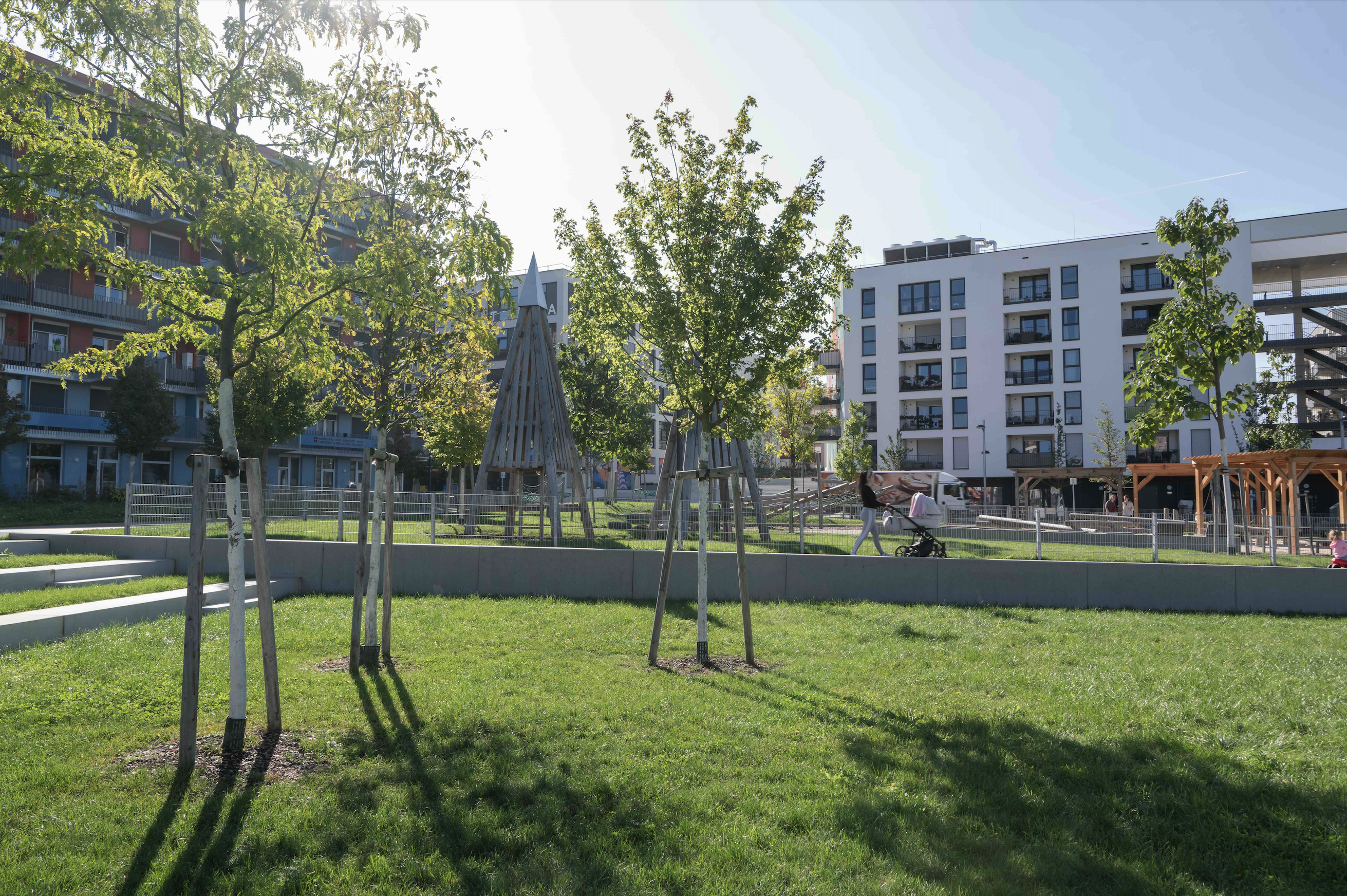 Elinor-Ostrom-Park: Parkfläche mit Bäumen, im Hintergrund eine Frau mit Kinderwagen und eine Hausanlage