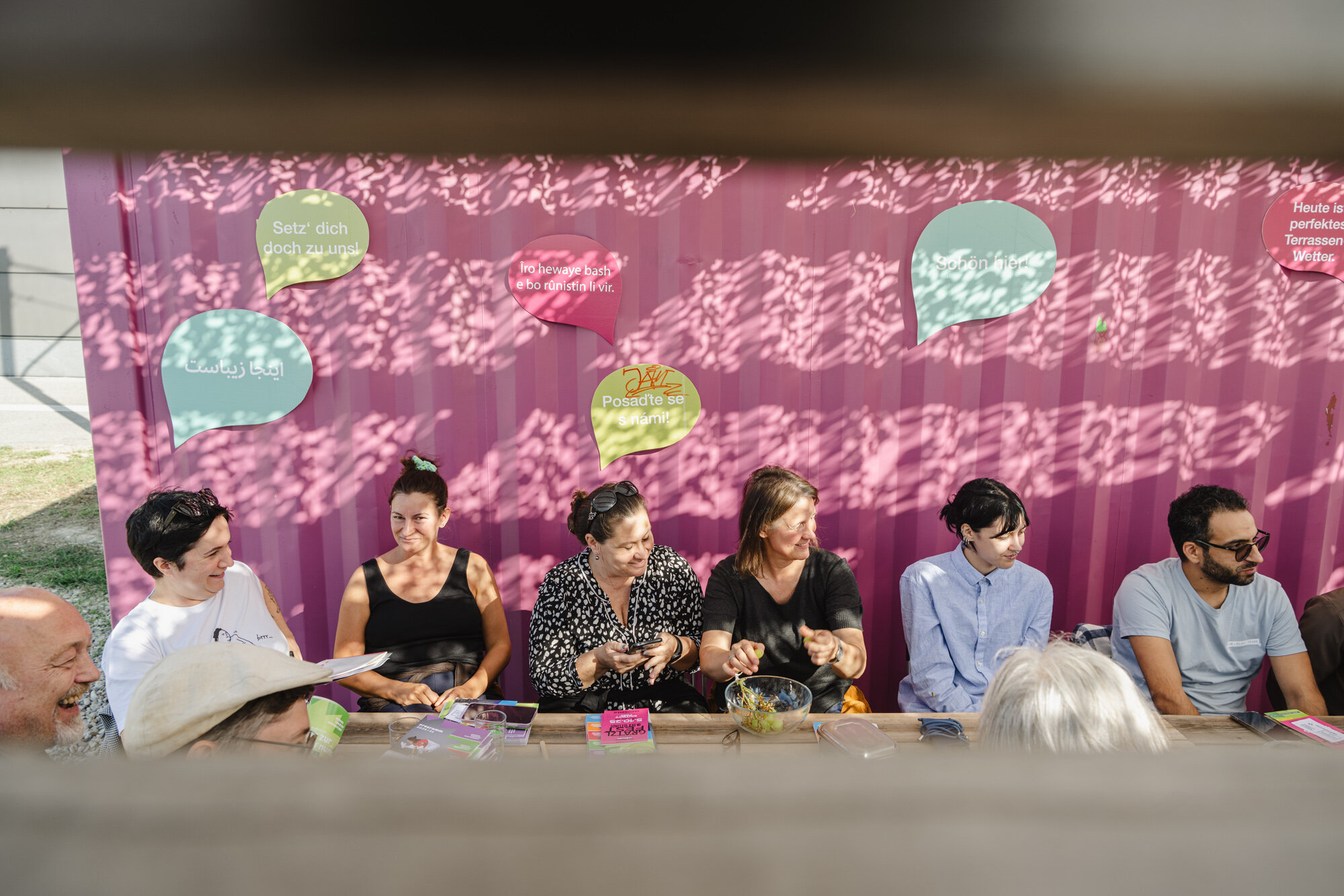 Menschen sitzen auf einer Bank. Hinter ihnen steht ein pinker Container