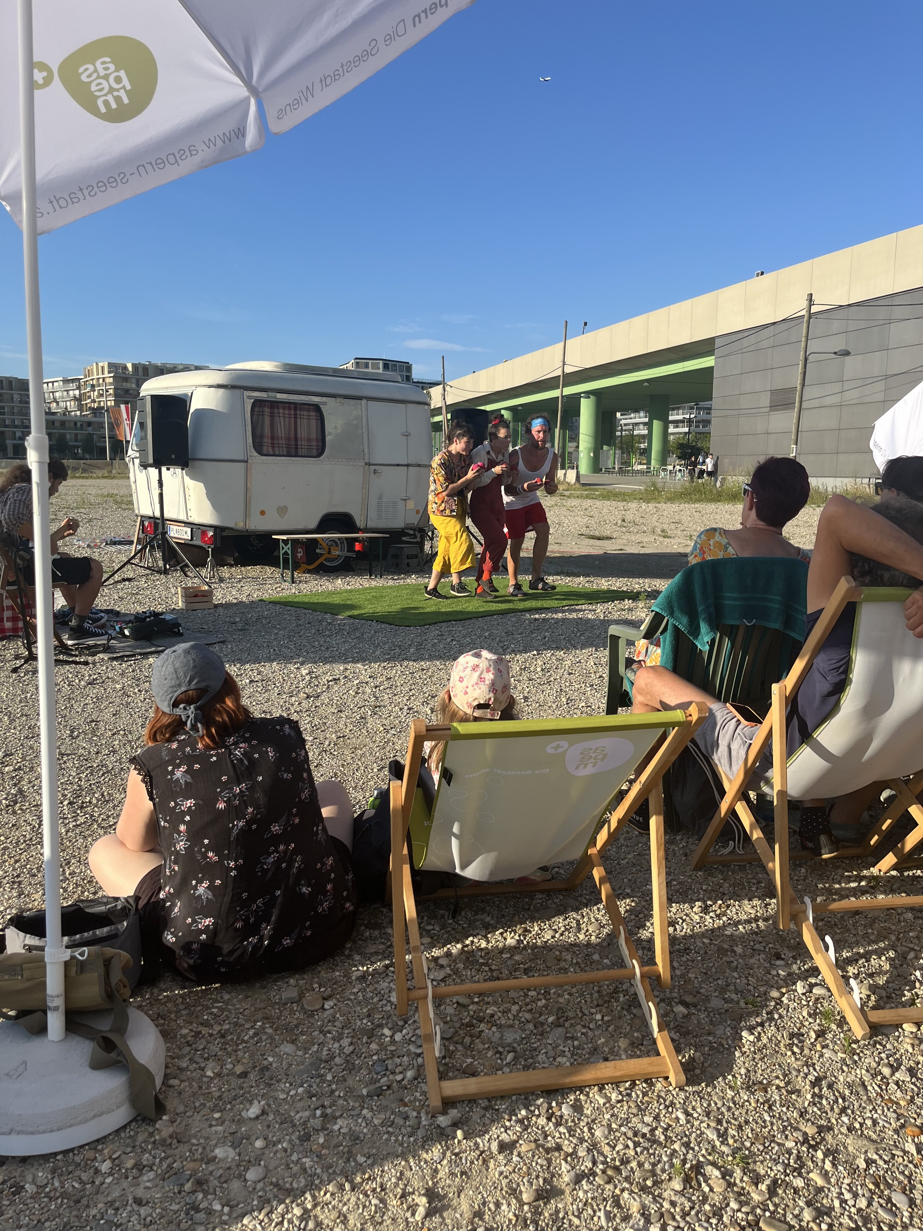 Drei Menschen führen ein Theater auf. Hinter ihnen steht ein silberner Wohnwagen