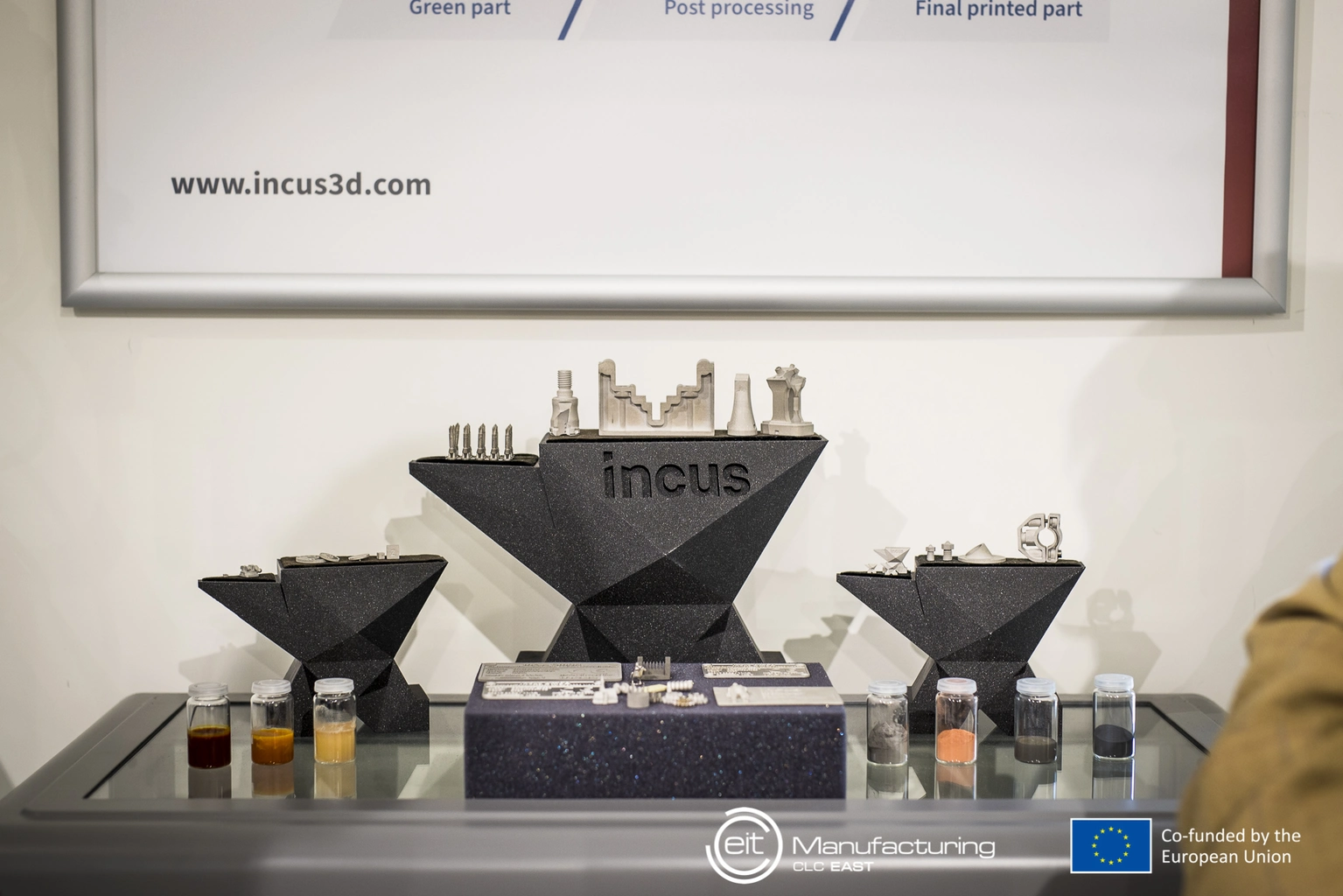 Eine Kommode mit Formen aus dem 3D-Drucker mit der Gravur Incus darauf, im Hintergrund ein White Board mit www.incus3d.com