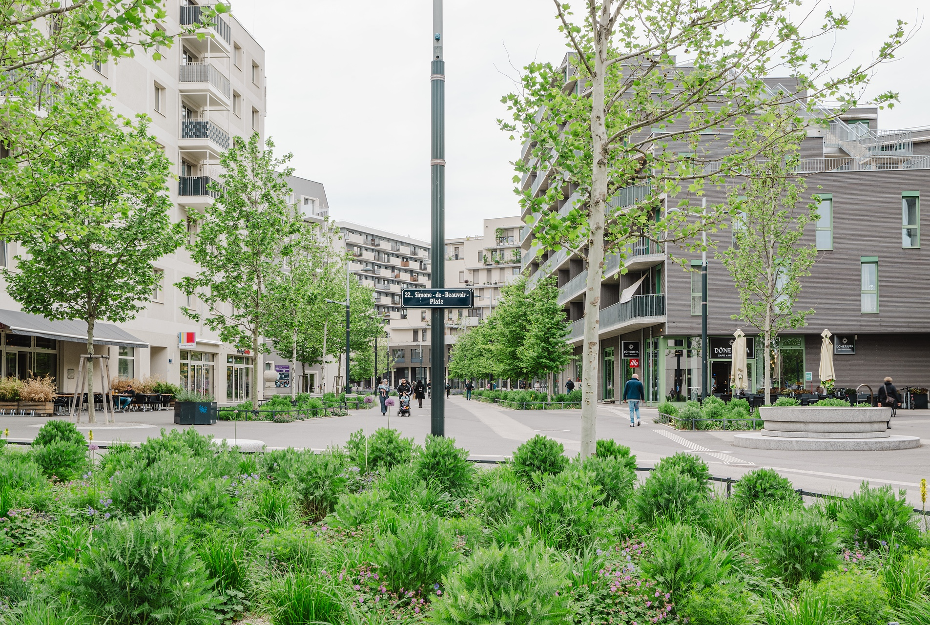 Straßenschild Simone de Beauvoir Platz mit Begrünung im Vordergrund und Häusern im Hintergrund