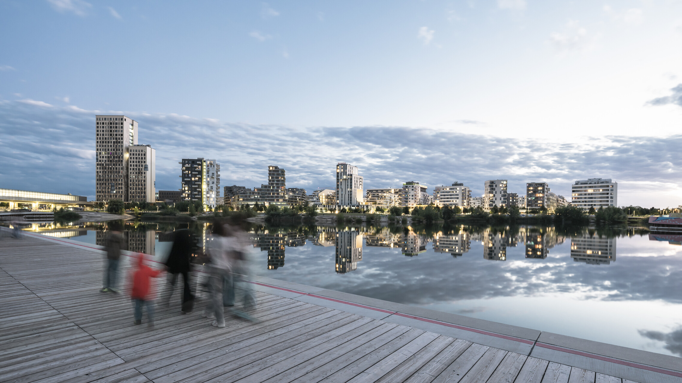 Blick von der Nordseite bei Dämmerung über den See auf die Skyeline der Seestadt mit verschwommenen Menschen im Vordergrund