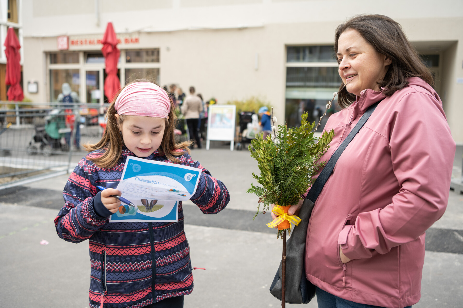 Kind mit Mutter im öffentlichen Raum, Kind schaut auf ein Blatt Papier in der Hand und versucht ein Rätsel zu lösen