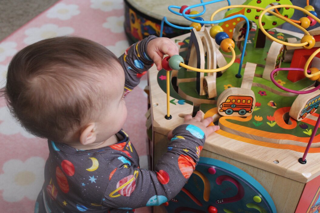 Baby mit dunklen Haaren spielt mit Motorikspielzeug, Baby ist von hinten zu sehen und trägt einen grauen Body mit Planeten darauf