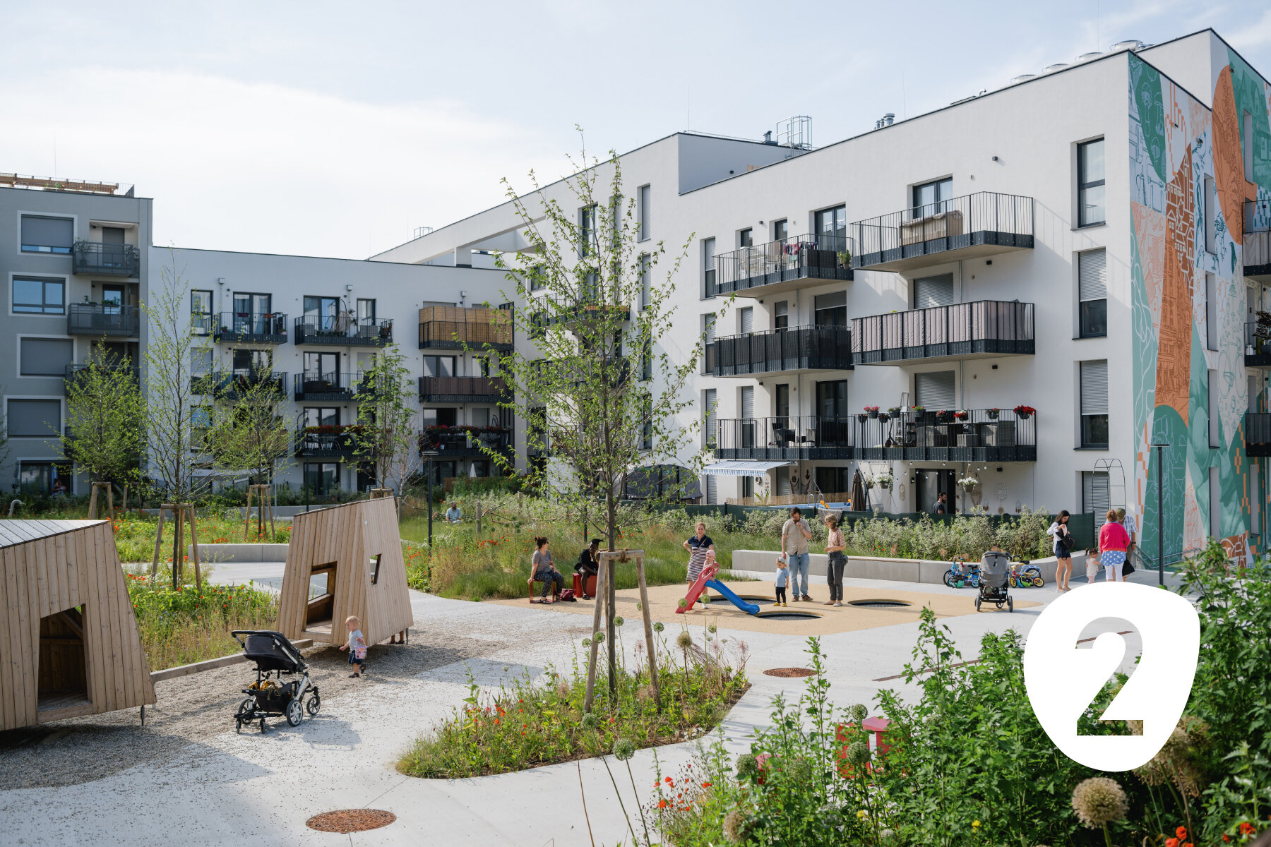 Menschen mit Kindern in einem großen begrünten Innenhof mit Holzspielplatz und hellen Gebäuden mit großen Balkonen