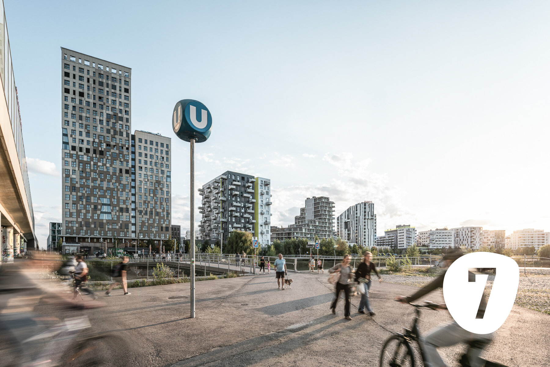 Menschen gehen oder fahren mit dem Rad vor einer U-Bahnstation am See mit moderner Skyline im Hintergrund
