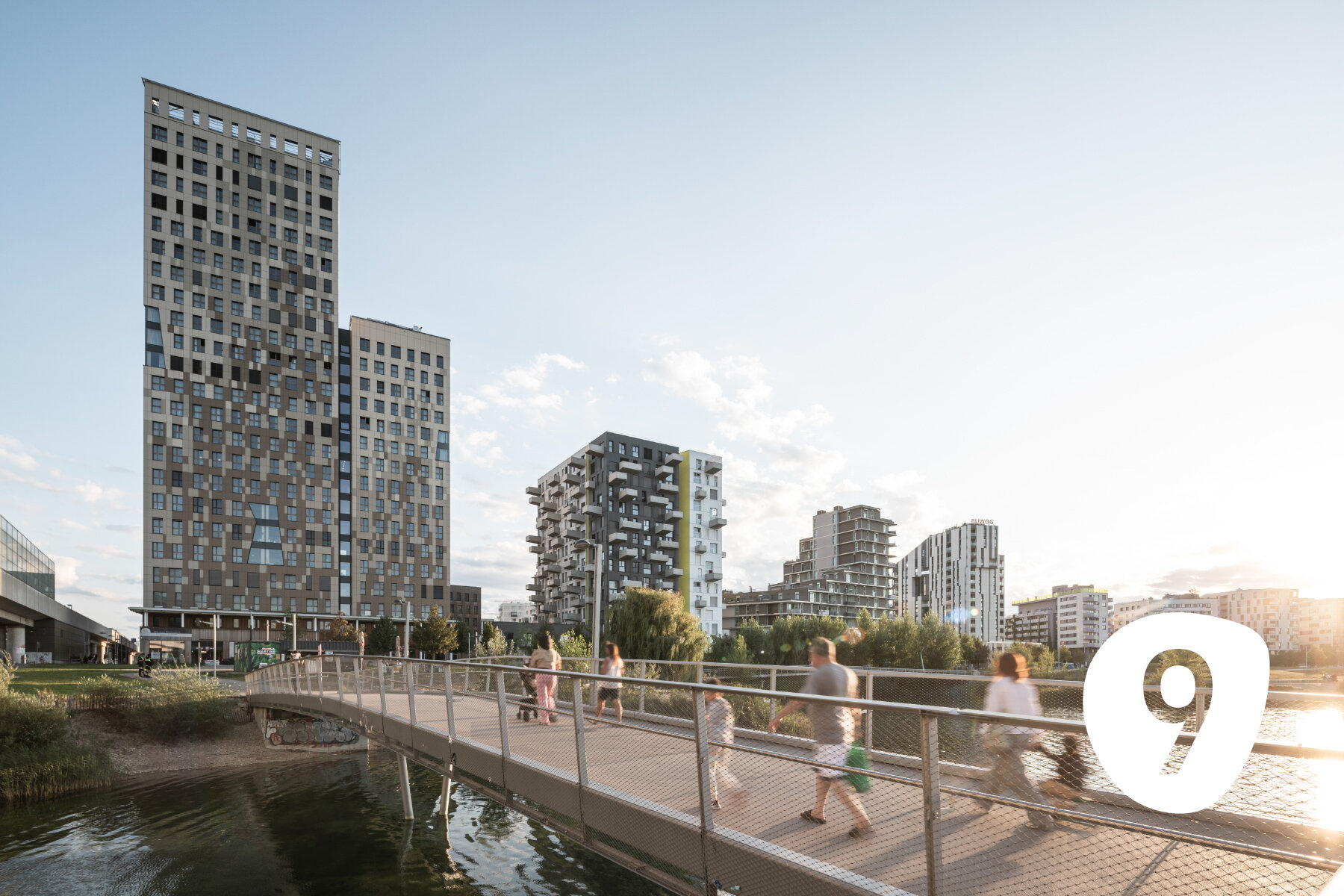 Menschen gehen über eine Brücke Richtung eines modernen Stadtteils, der an einem See liegt