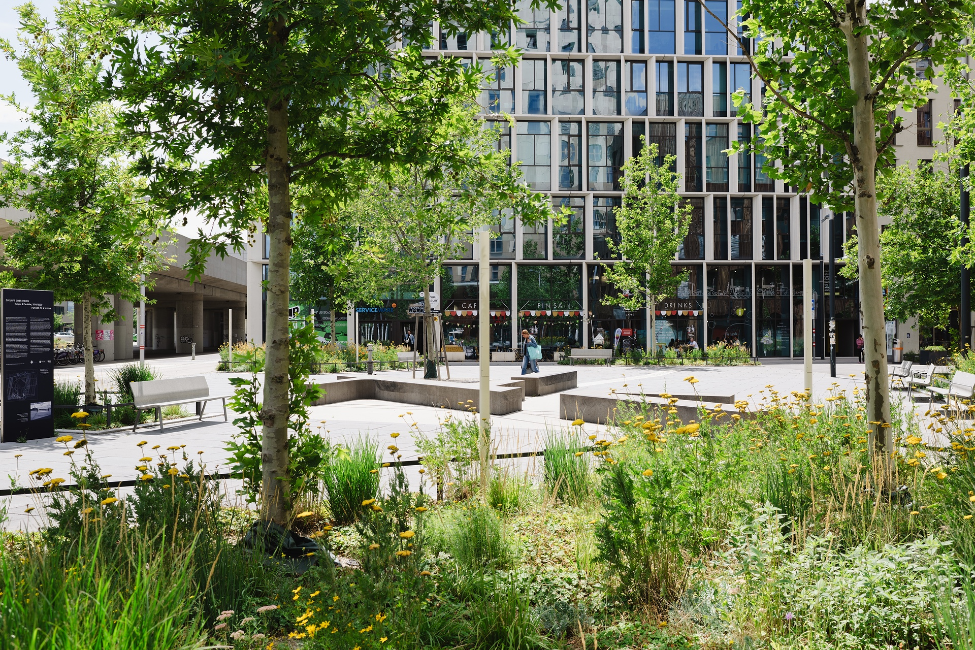 Bild eines grünen Platzes mit angedeuteten Menschen und Gebäuden im Hintergrund. 