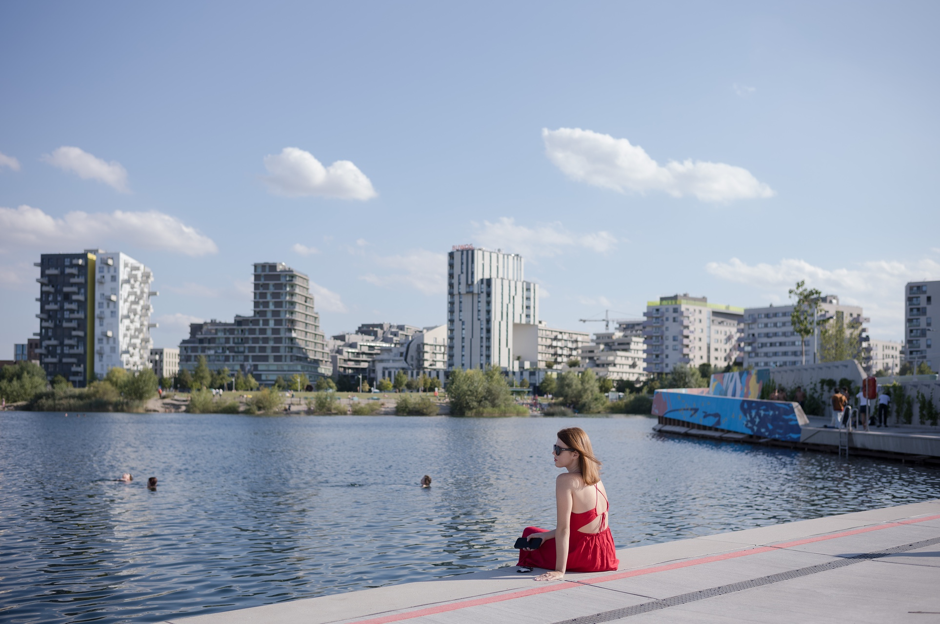 Frau in rotem Kleid sitzt am Seeufer und schaut in Richtung Seestadt-Skyline.