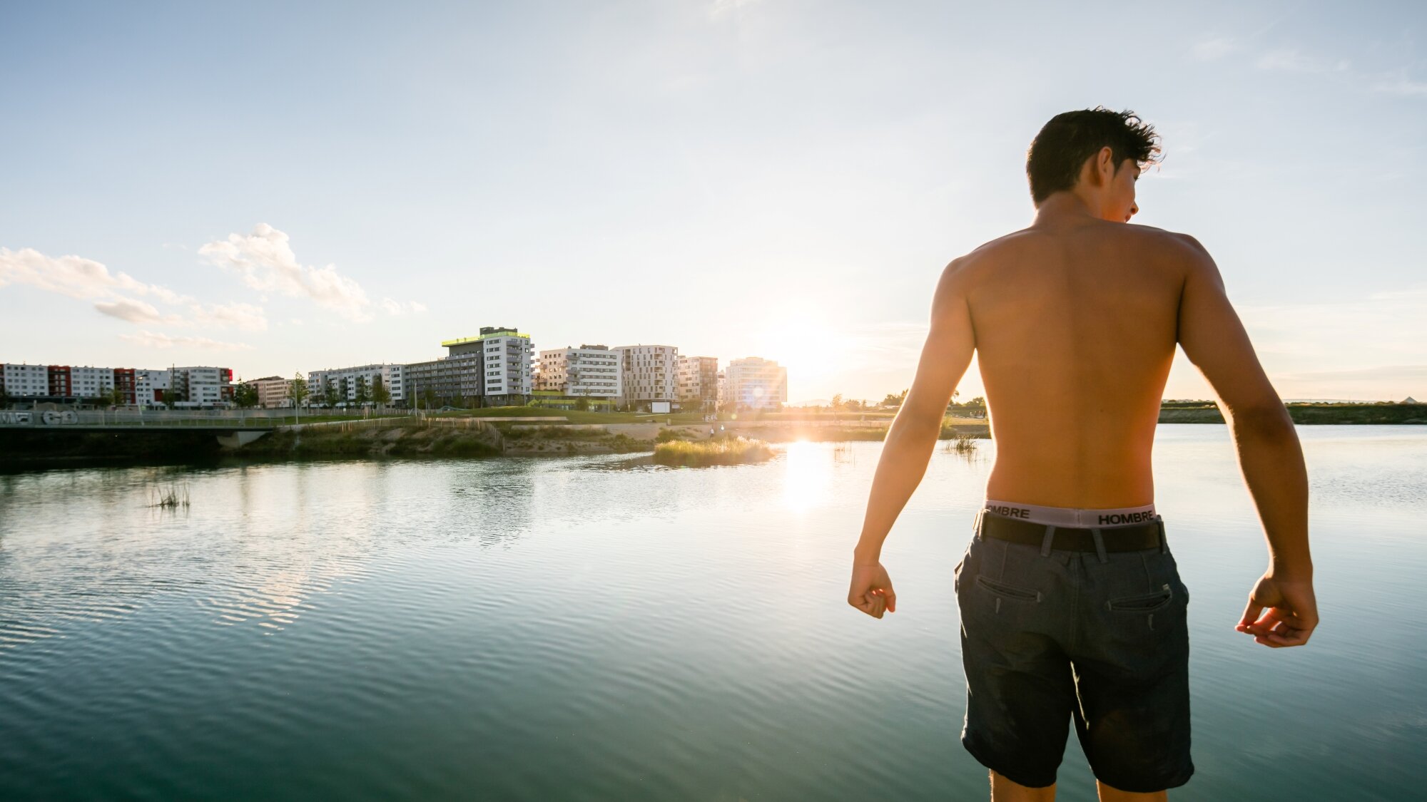 junger Mann in Badehose steht an einem See vor der Skyline einer modernen Stadt