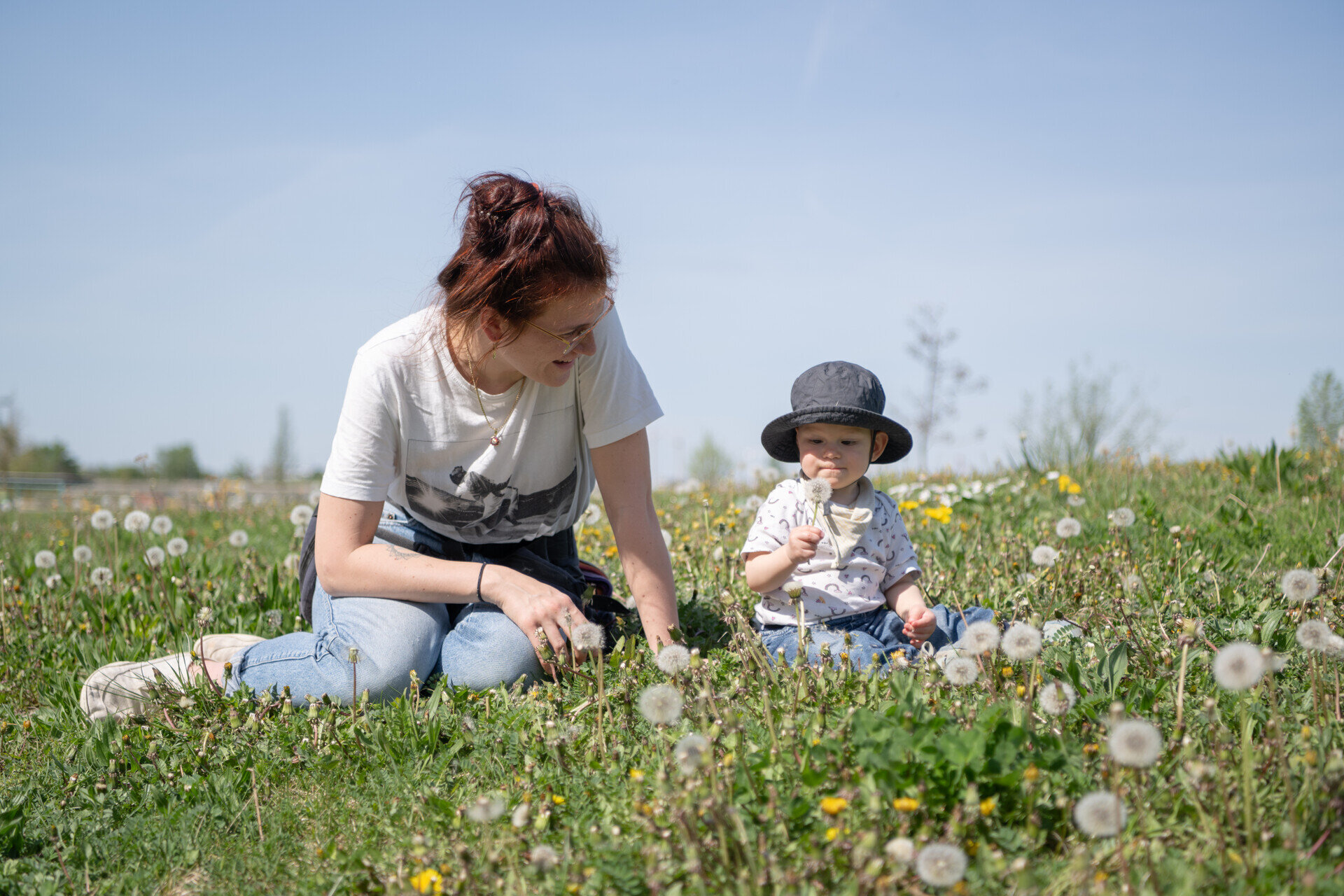 Frau und Kleinkind sitzen auf einer blühenden Wiese mit Löwenzahn, beide wirken entspannt und genießen die Sonne.