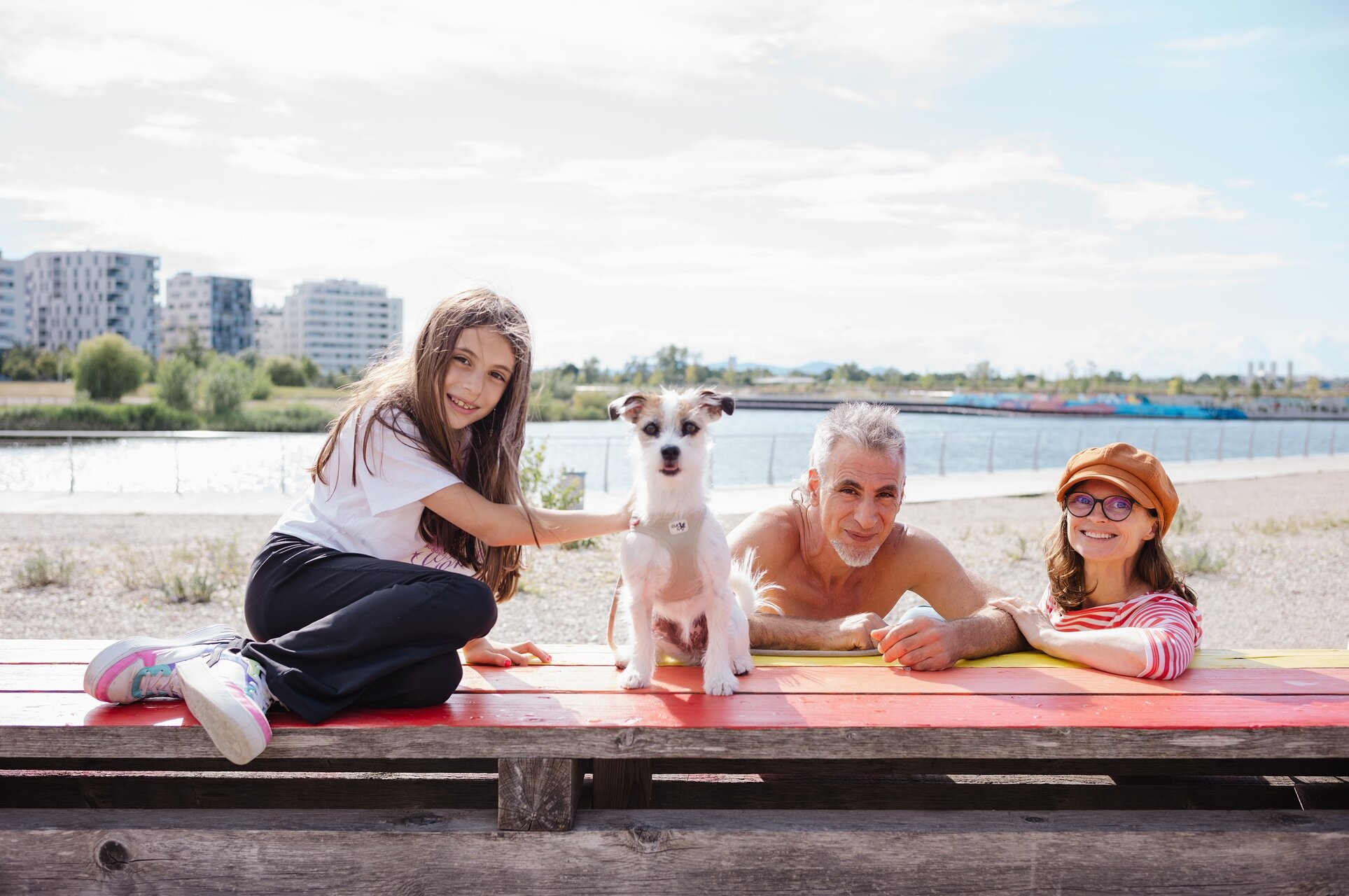 Zwei Kinder, ein älterer Mann und ein Hund sitzen gemeinsam auf einer bunten Bank am Ufer des asperner Sees, im Hintergrund Wasser und moderne Wohnhäuser.