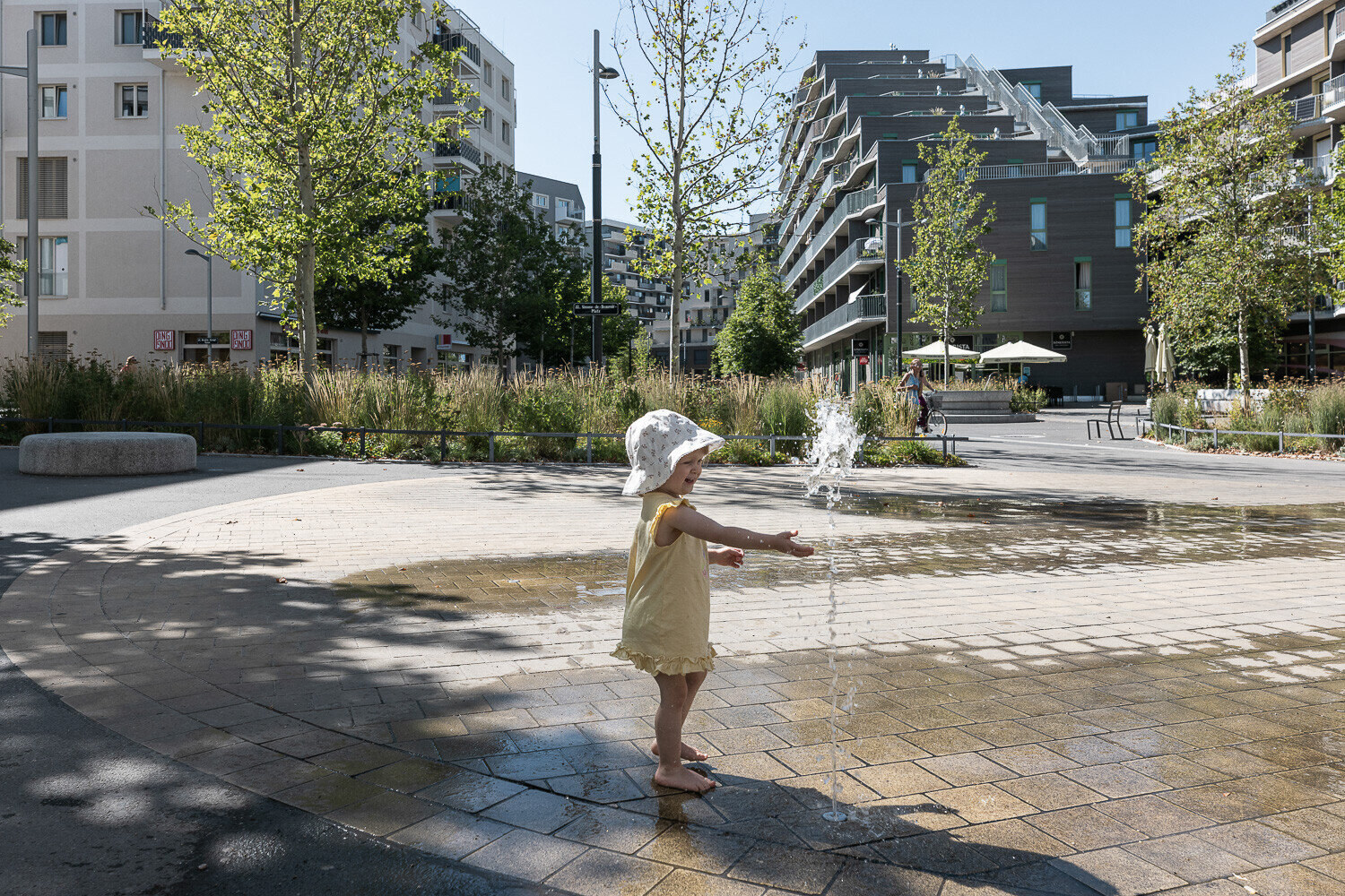 Kleines Kind spielt lachend an einem Wasserspielplatz in der Seestadt, umgeben von modernen Wohnhäusern und junger Begrünung.