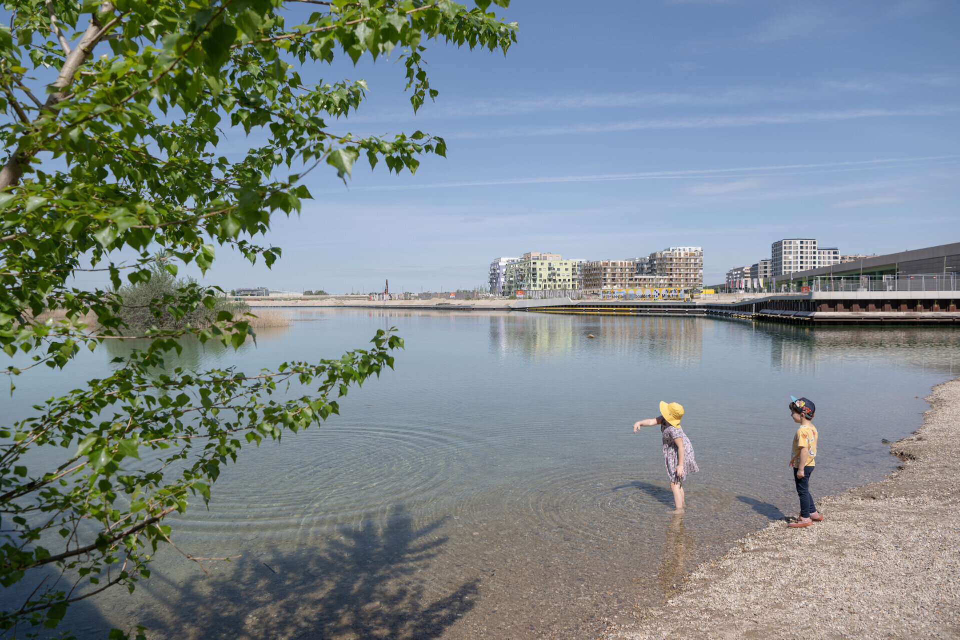 Zwei Kinder spielen am Ufer des asperner Sees, eines steht mit den Füßen im Wasser, während im Hintergrund moderne Wohngebäude zu sehen sind.
