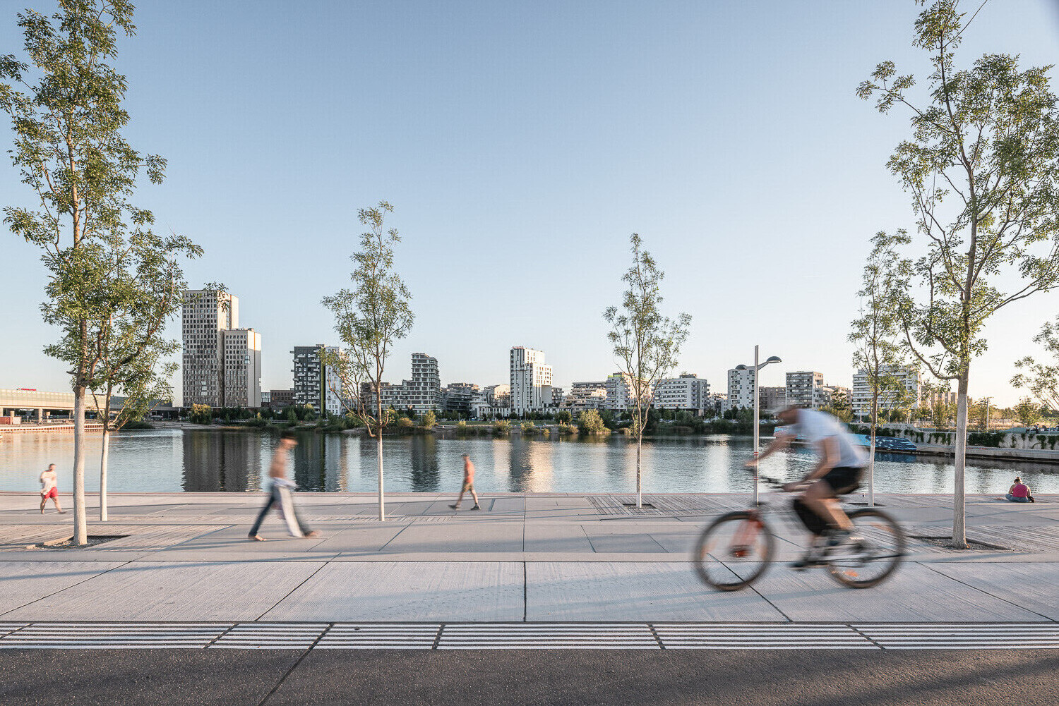 Drei Personen spazieren auf der Seestadtpromenade, eine Person ist auf dem Rad zu sehen, alle sind durch die Bewegung verschwommen. Im Hintgrund scharf der See und die Skyline der Seestadt