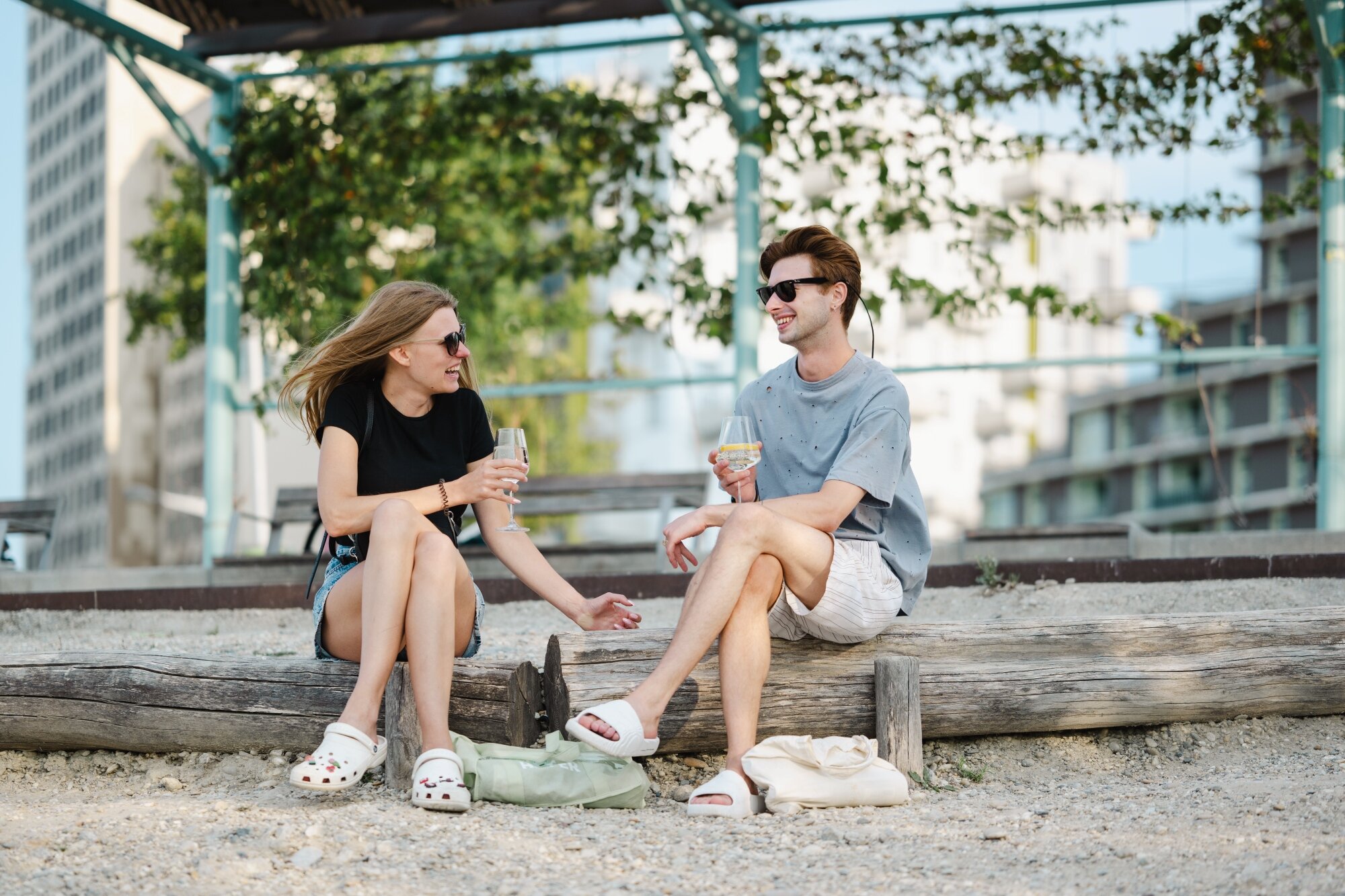 Frau und Mann sitzen auf Holzstamm am Kiestrand unter einer Pergola mit Weingläsern vor einer urbanen Skyline