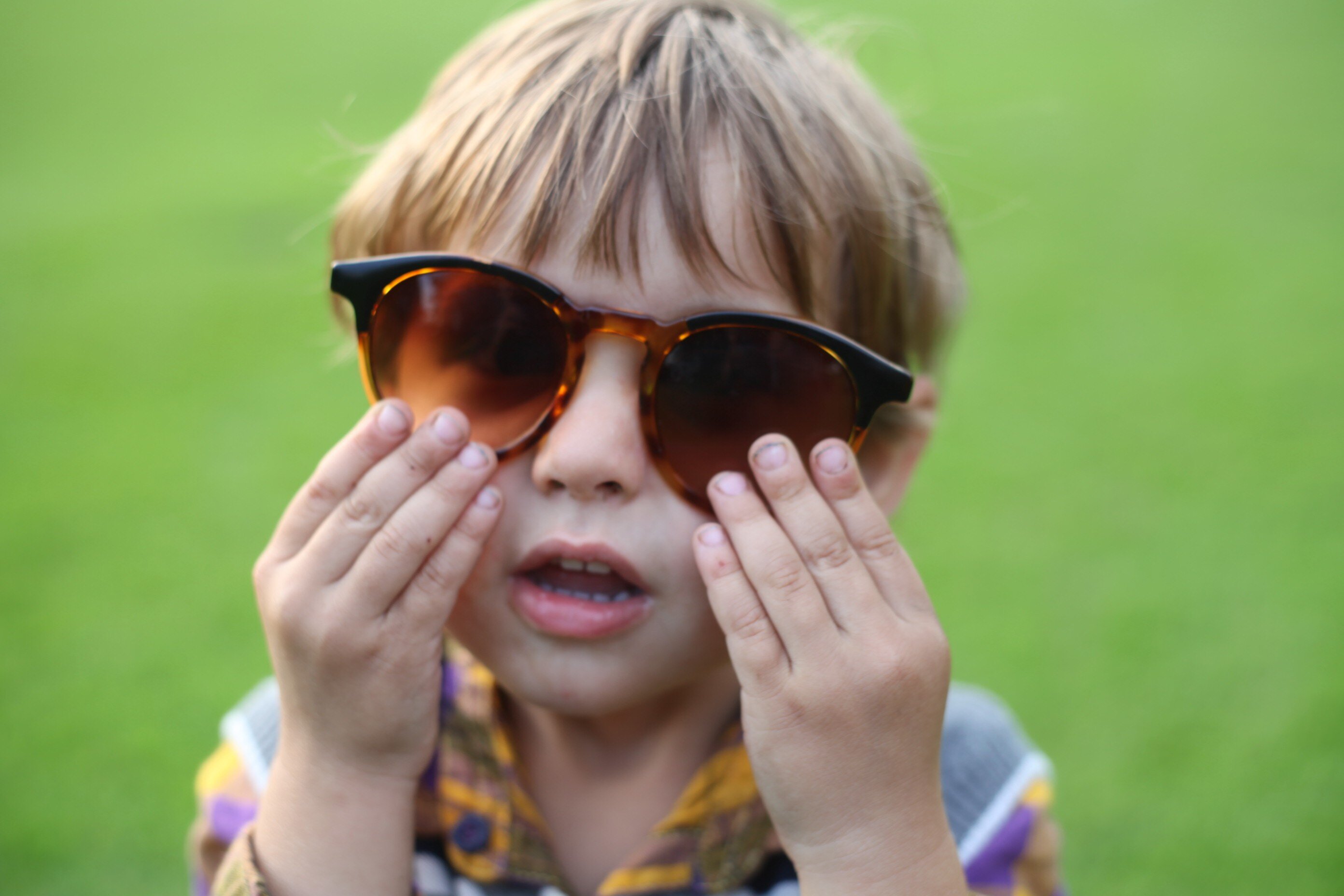 Kind mit Sonnenbrille auf einer Wiese Kind mit brauner Sonnenbrille und bunt kariertem Hemd steht auf einer Wiese und hält sich die Hände ans Gesicht.
