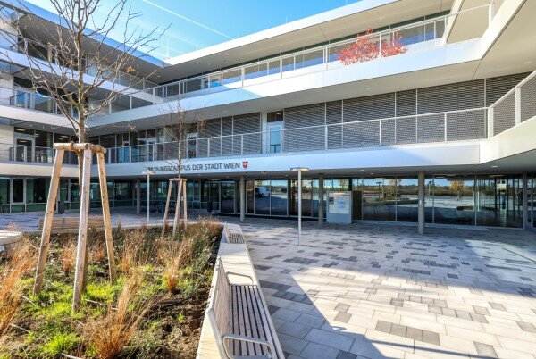 Bildungscampus Liselotte-Hansen-Schmidt Modernes Schulgebäude mit Glasfront und Innenhof, Sonnenlicht und junge bepflanzte Bäume im Vordergrund.