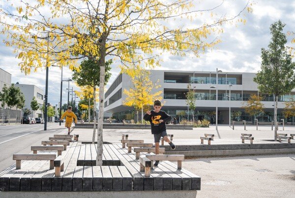 Bundesgymnasium Seestadt Kinder laufen und spielen auf einer modernen Platzanlage mit Bänken und jungen Bäumen vor einem Schulgebäude.