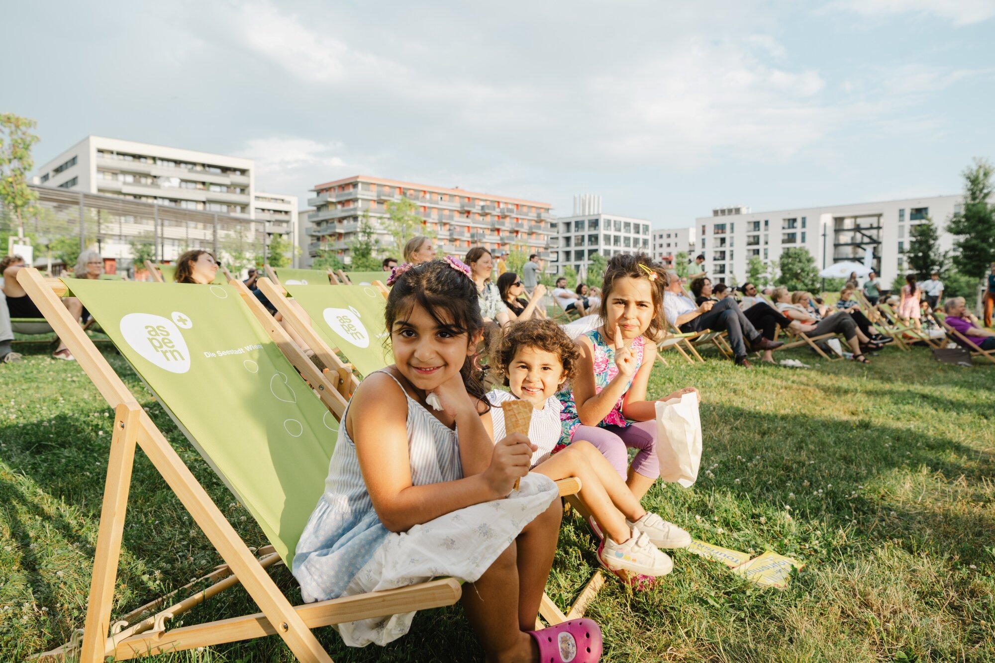 Kinder sitzen in einem Liegstuhl auf einer großen Wiese mit vielen Menschen und modernen Wohnhäusern im Hintergrund