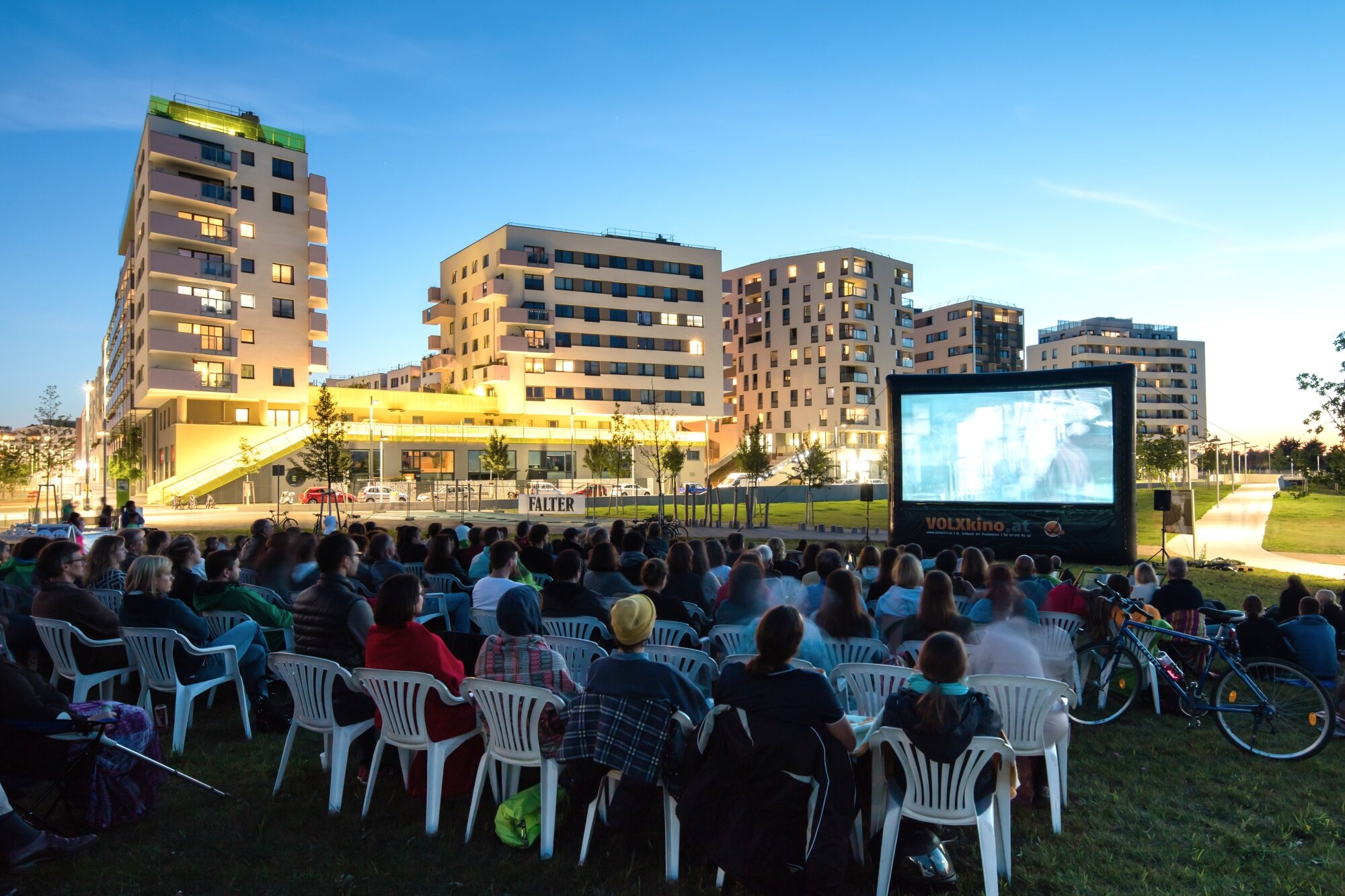 Open-Air-Kino - Freizeitprogramm in der Seestadt viele Menschen sitzen im einem großen Park am Abend beim Open-Air-Kino mit modernen Gebäuden im Hintergund