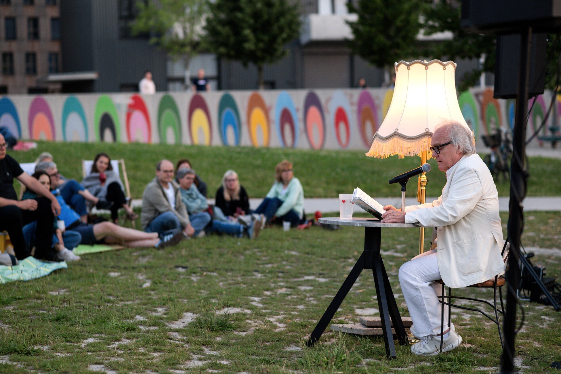 Mondscheinlesung der Buchhandlung Seeseiten Mann in weißem Anzug liest im Park aus einem Buch mit aniker Stehlampe und bemalter Wand