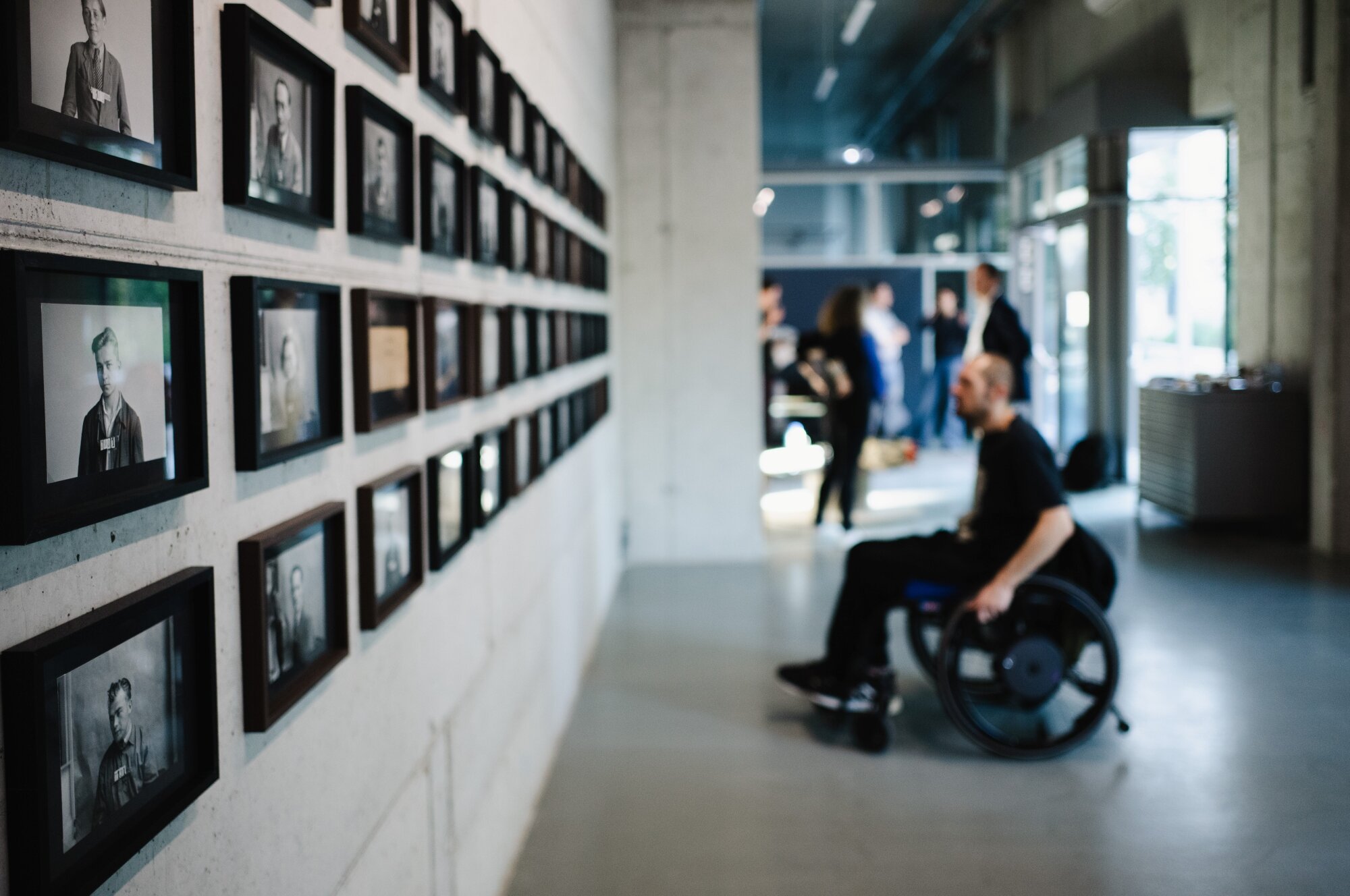 Bilder von Maria Zisser im LLLLLL Mann im Rollstuhl betrachtet eine Fotoausstellung in einem modernen Kunstraum mit weiteren Menschen
