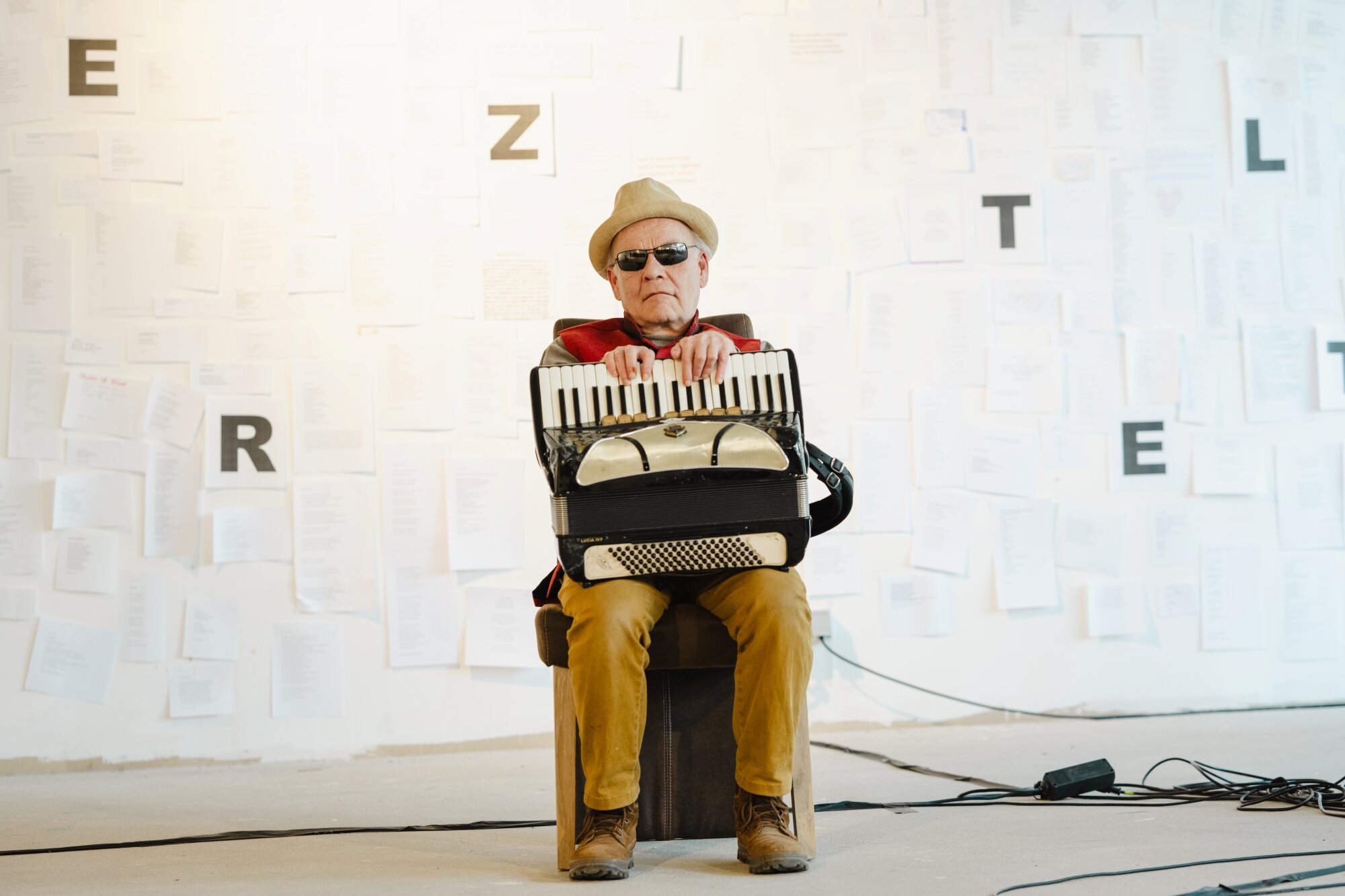 Otto Lechner bei LYRIK ON LINE älterer Mann mit Sonnenbrille und Akkordeon sitzt auf einem Stuhl vor einer weißen Wand mit Buchstaben