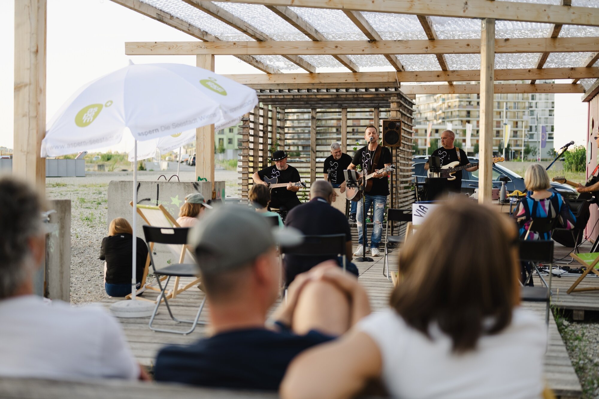 eine Band spielt auf einer Holzterrasse mit Pergola vor einem leeren Baufeld und bunten Wohnhäusern