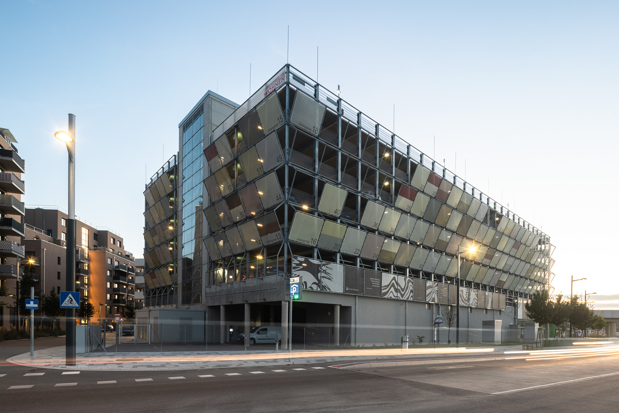 futuristisches Gebäude mit Bannern vor den Parkflächen der mehrstöckigen Garage mit Kulturfläche im Erdgeschoß