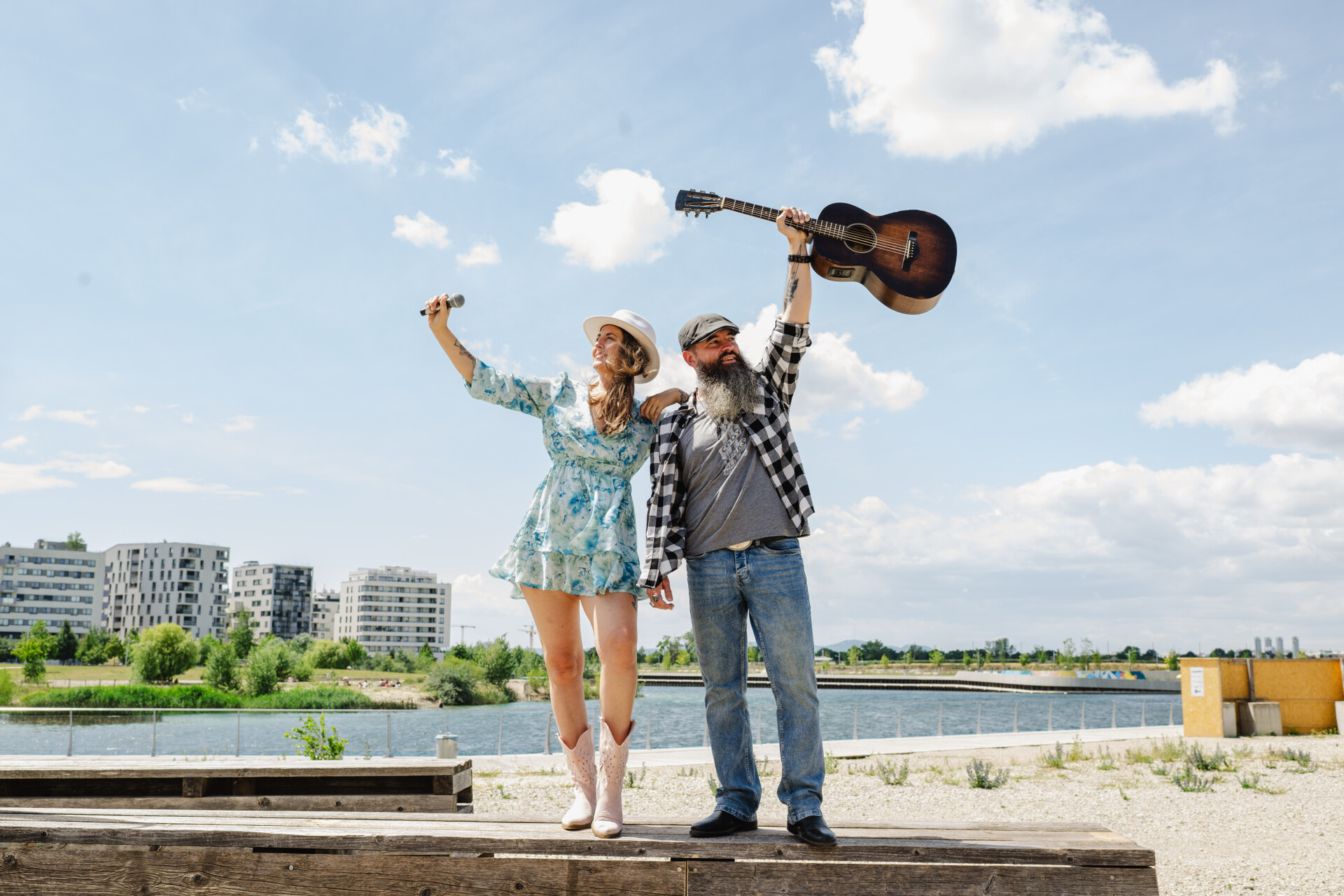 Zwei Musiker - Frau und Mann - halten Mikrofon und Gitarre in die Luft. Im Hintergrund See. 