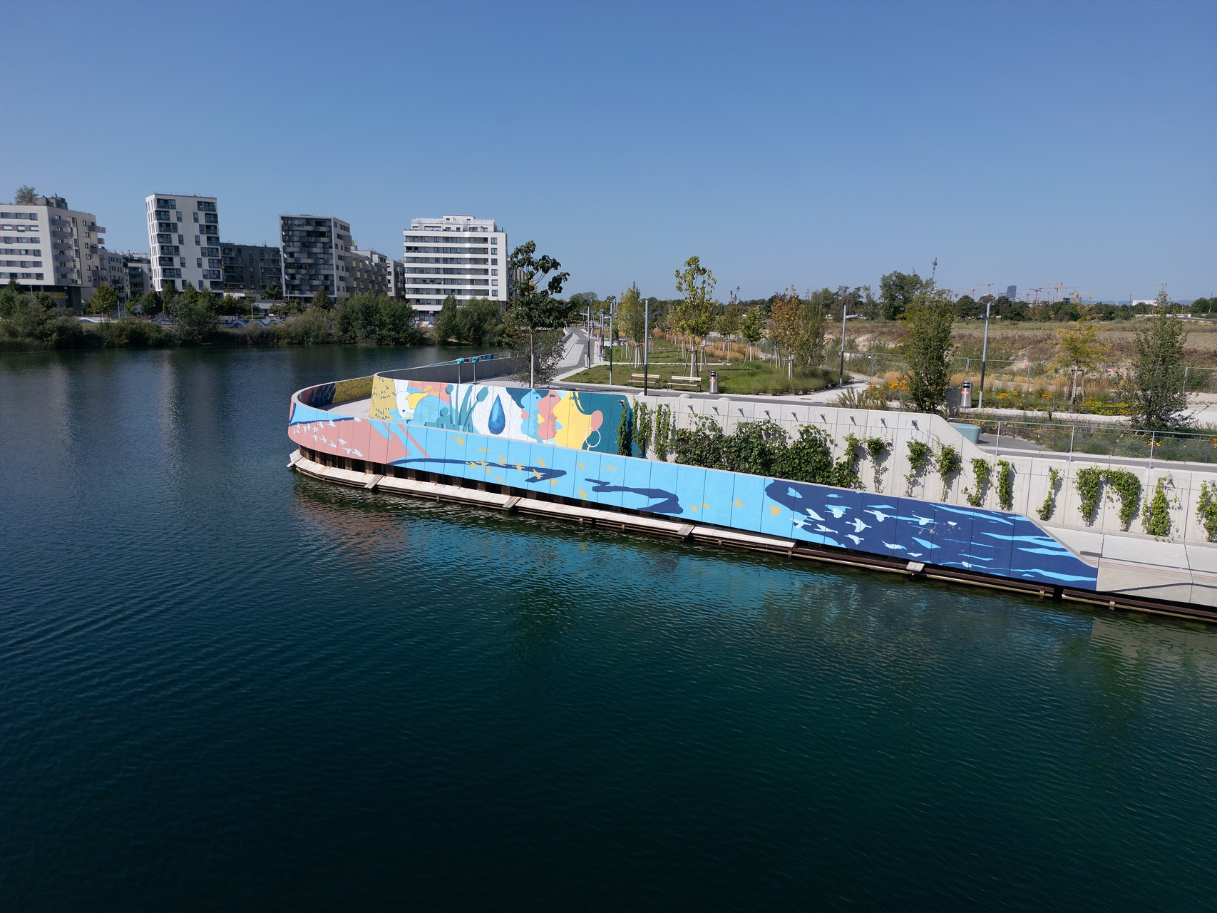 Blaues Mural an der Wand einer Promenade, davor ein See, dahinter Grünflächen mit Bäumen sowie auf der linken Seite Häuser