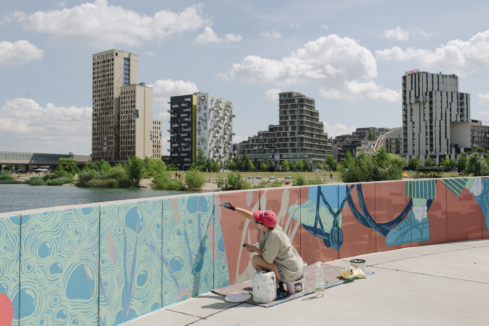 Künstler*in bemalt die Wand an der Promenade mit bunden Farben, im Hintergrund ein See und dahinter ein Park sowie Gebäude