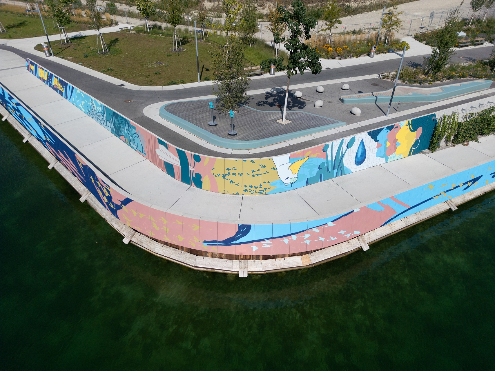 Mural mit bunten Frauen, einer Ente und unterschiedlichen Farben an der Wand einer Promenade, dahinter Grünflächen mit Bäumen