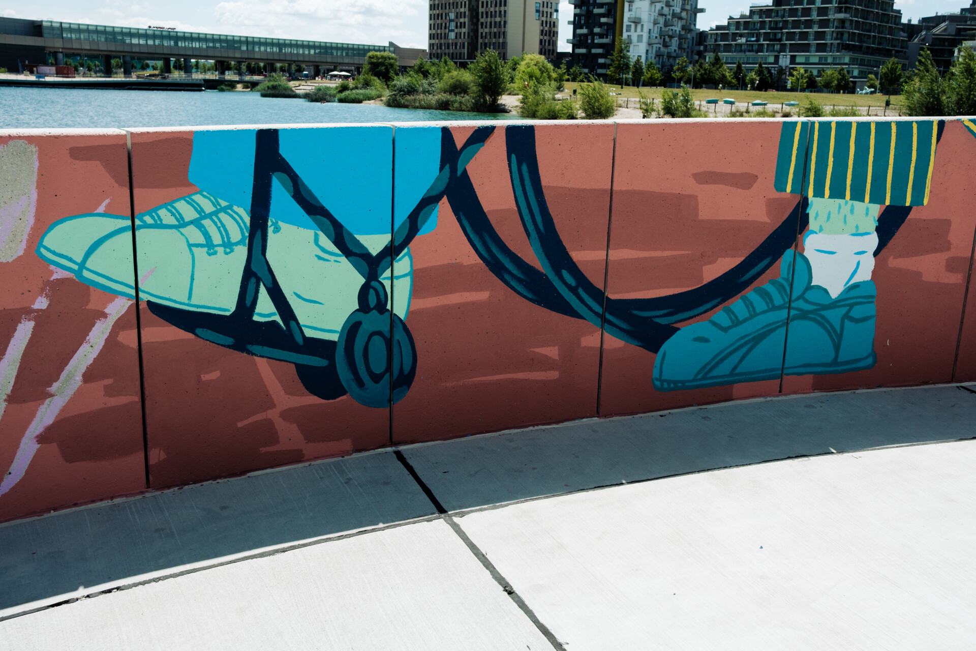 Mural an der Promenadenwand: Nut der untere Teil eines Rollstuhles mit Füßen des im Rollstuhl sitzenden und einem Fuß der Person, die schiebt, dahinter der See, die U-Bahn-Station, ein Park und Gebäude