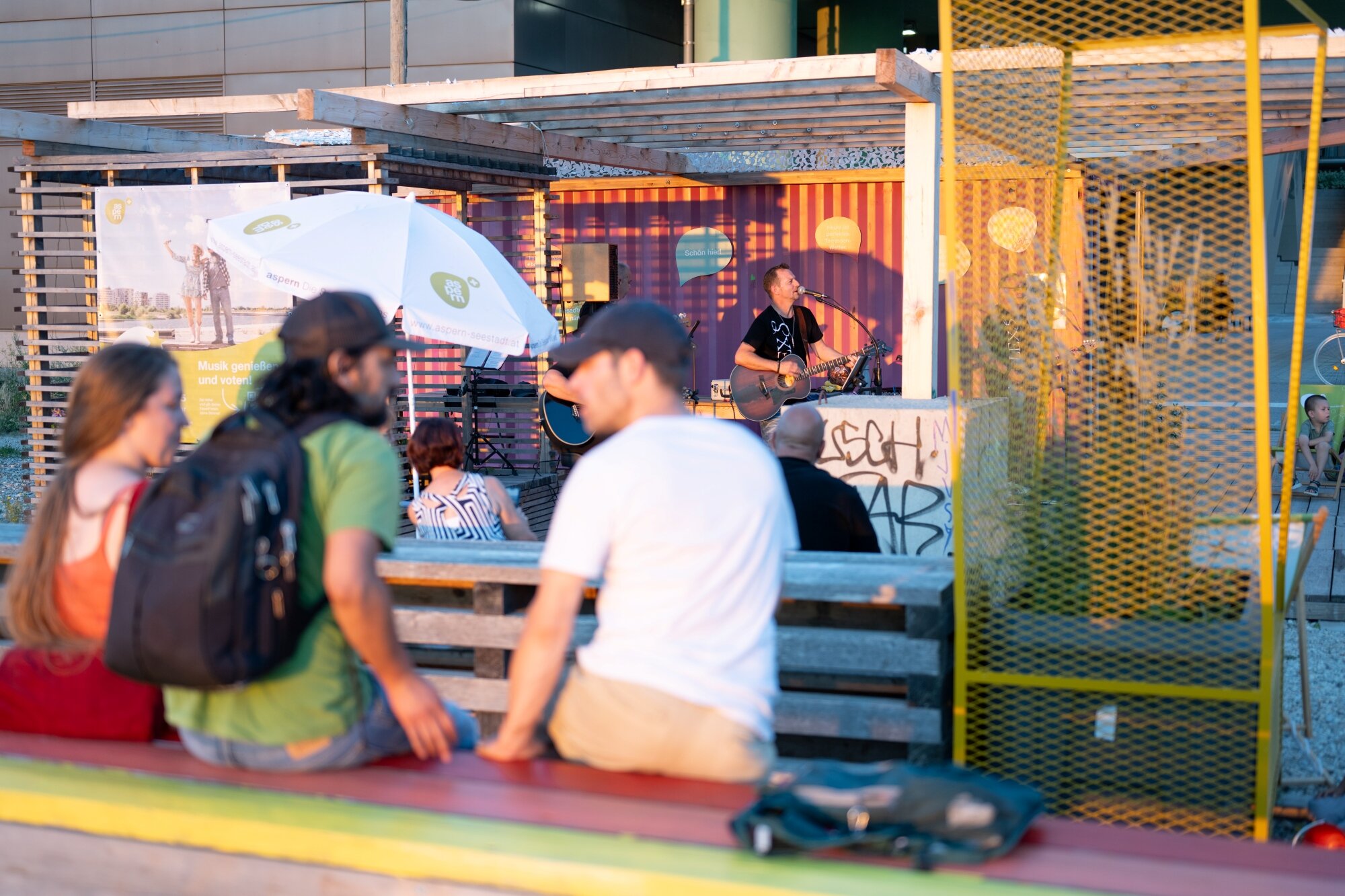 temporäre Nachbarschaftsterrasse am See Menschen sitzen auf bunten Holzmöbeln bei einem Konzert vor einem bunten Container auf einer Terrasse
