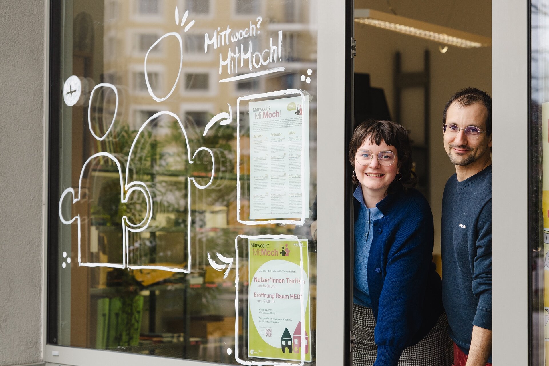 STM Leben Zwei Personen lächeln aus der Tür eines Nachbarschaftsraums in der Seestadt, auf der Glasscheibe sind Plakate und Zeichnungen für die Initiative „MitMoCH“ angebracht.