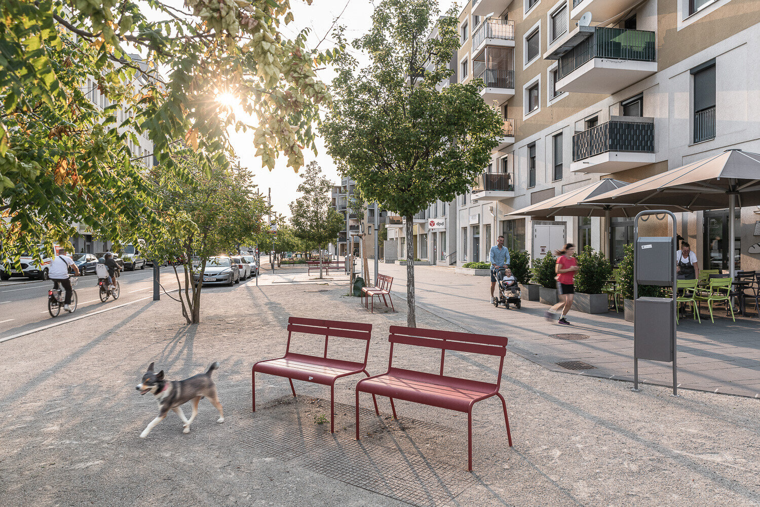 elebte Straße mit Geschäften, Cafés und roten Bänken in der Seestadt, Menschen spazieren, fahren Rad oder sitzen im Freien, die Sonne steht tief.