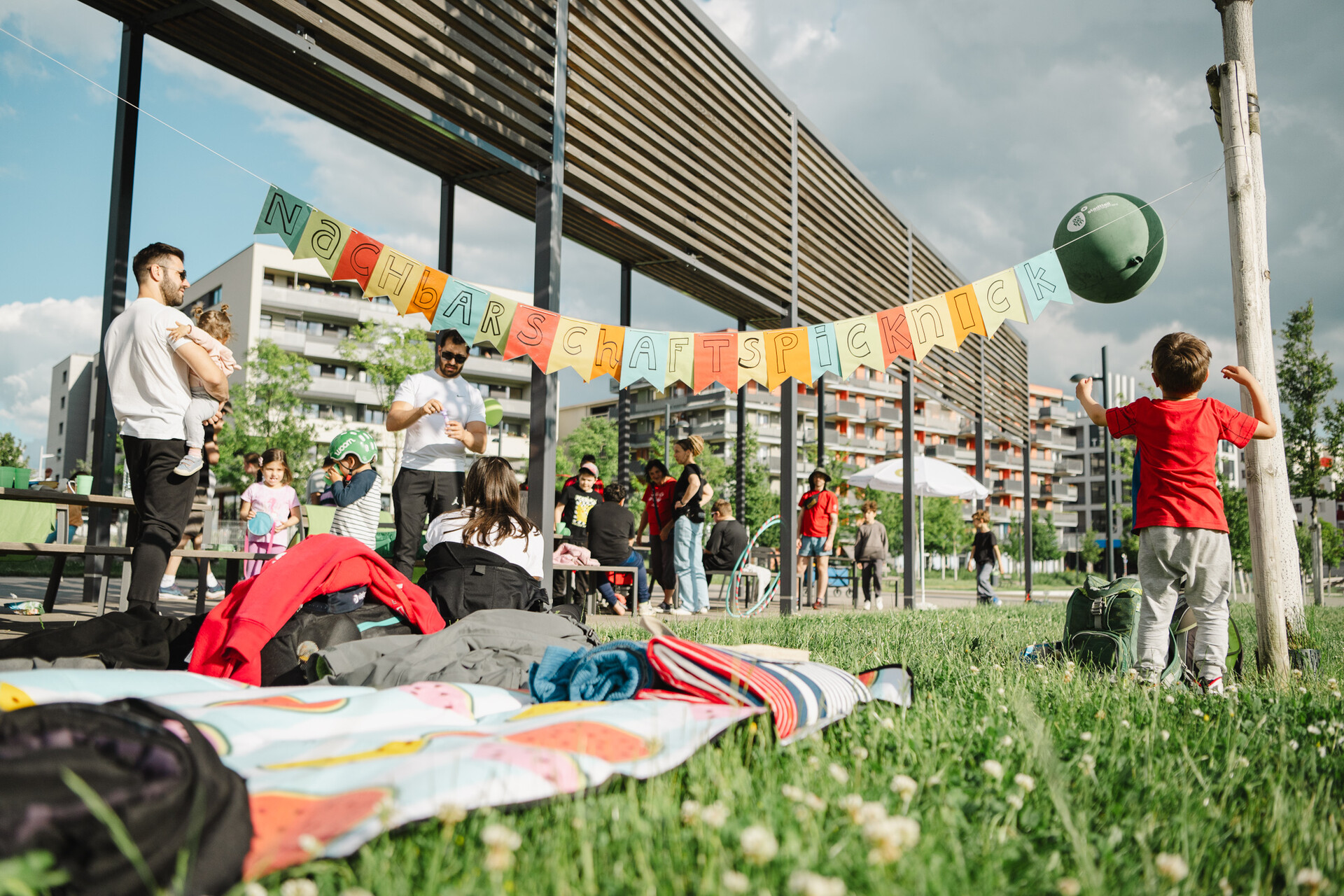 Picknickdecke auf der Wiese, mehrere Personen und ein Junge der einen Ball wirft, in der Mitte eine Girlande mit der Aufschrift Nachbarschaftspicknick