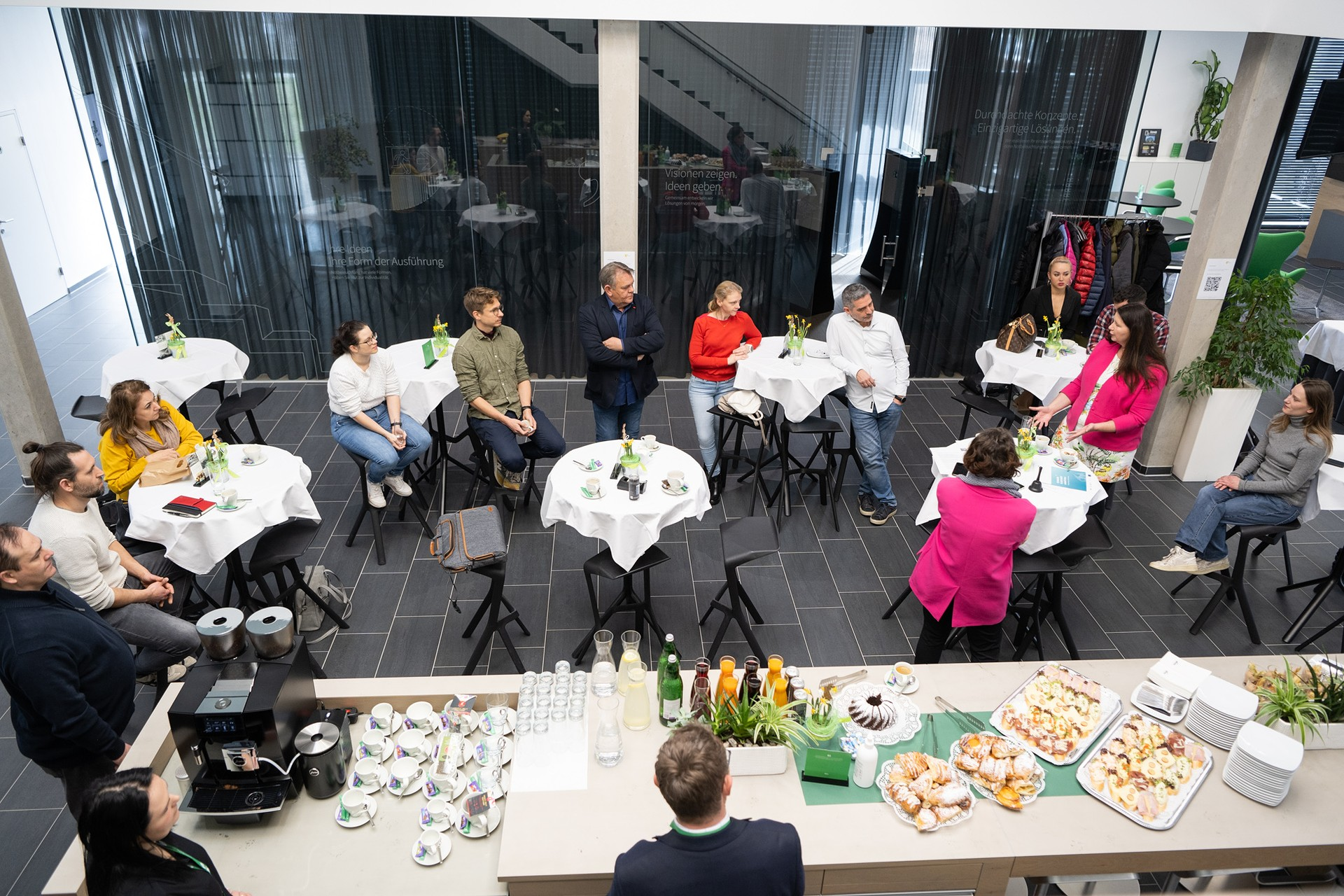 Eine Gruppe von Menschen steht und sitzt  um Stehtische, eine Frau in einem rosa Blazer spricht. Es stehen Frühstück und Getränke bereit. 