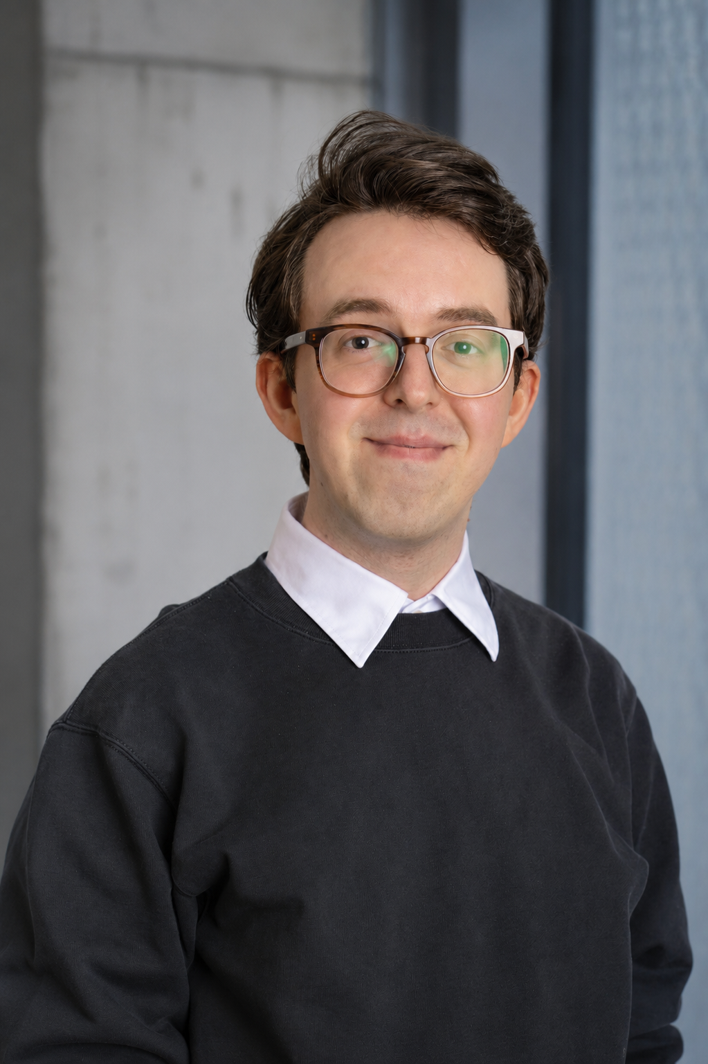 Portrait eines Mannes mit Brille und schwarzem Pullover vor einer grauen Betonwand