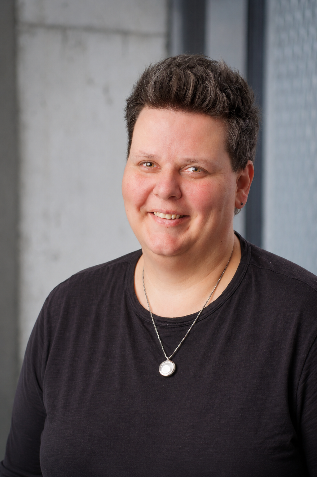 Portraitfoto einer Frau mit kurzen Haaren und schwarzem T-Shirt vor einer grauen Betonwand