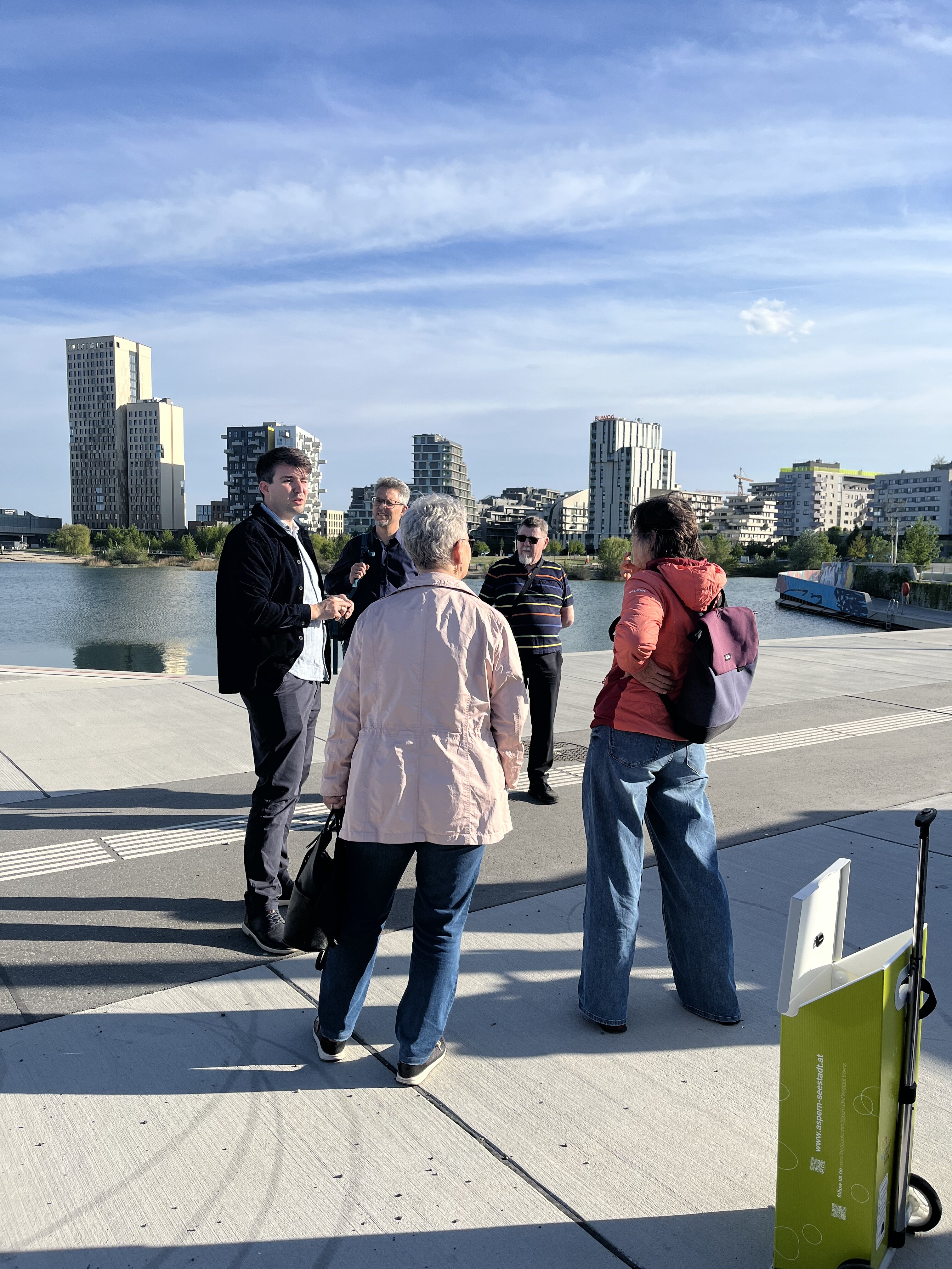 Menschen stehen am DOCK Seestadt und hören Tour-Guide zu, See und Skyline im  Hintergrund zu sehen