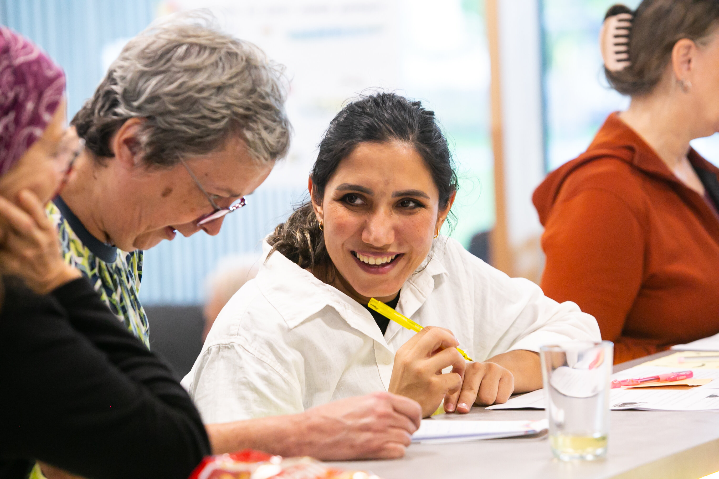 Drei Frauen sind zu sehen, sitzend an einem Tisch, sie schreiben etwas, eine Frau schaut dabei auf und lacht