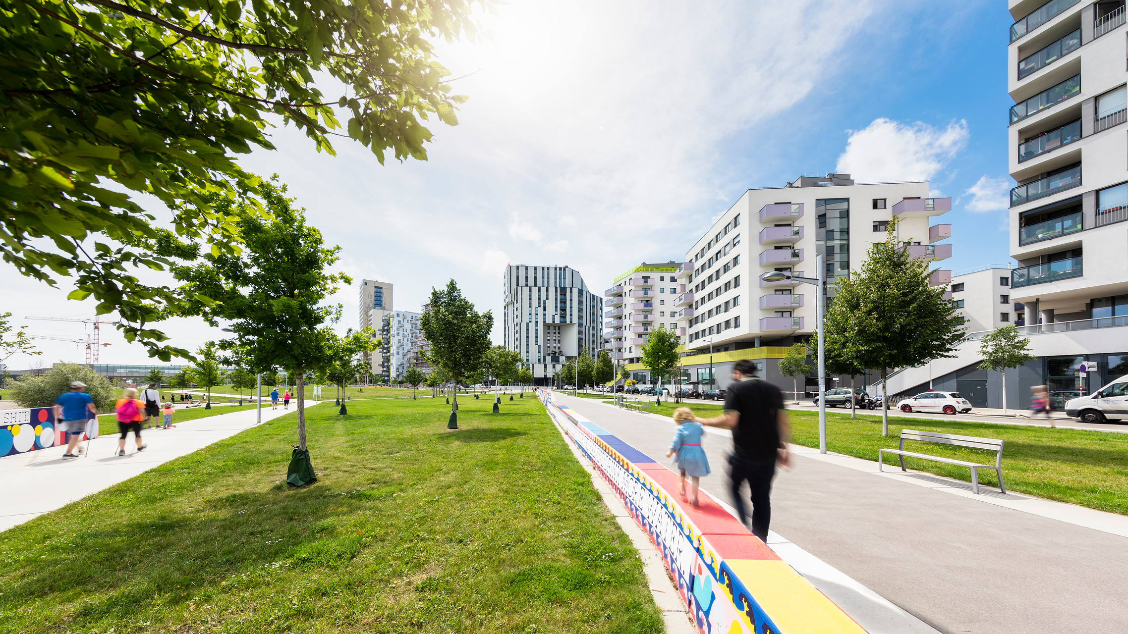 Blick auf einen Park mit Menschen, die spazieren, es scheint die Sonne, ein Mädchen balanciert an der Hand eines Mannes auf einer niedrigen Mauer