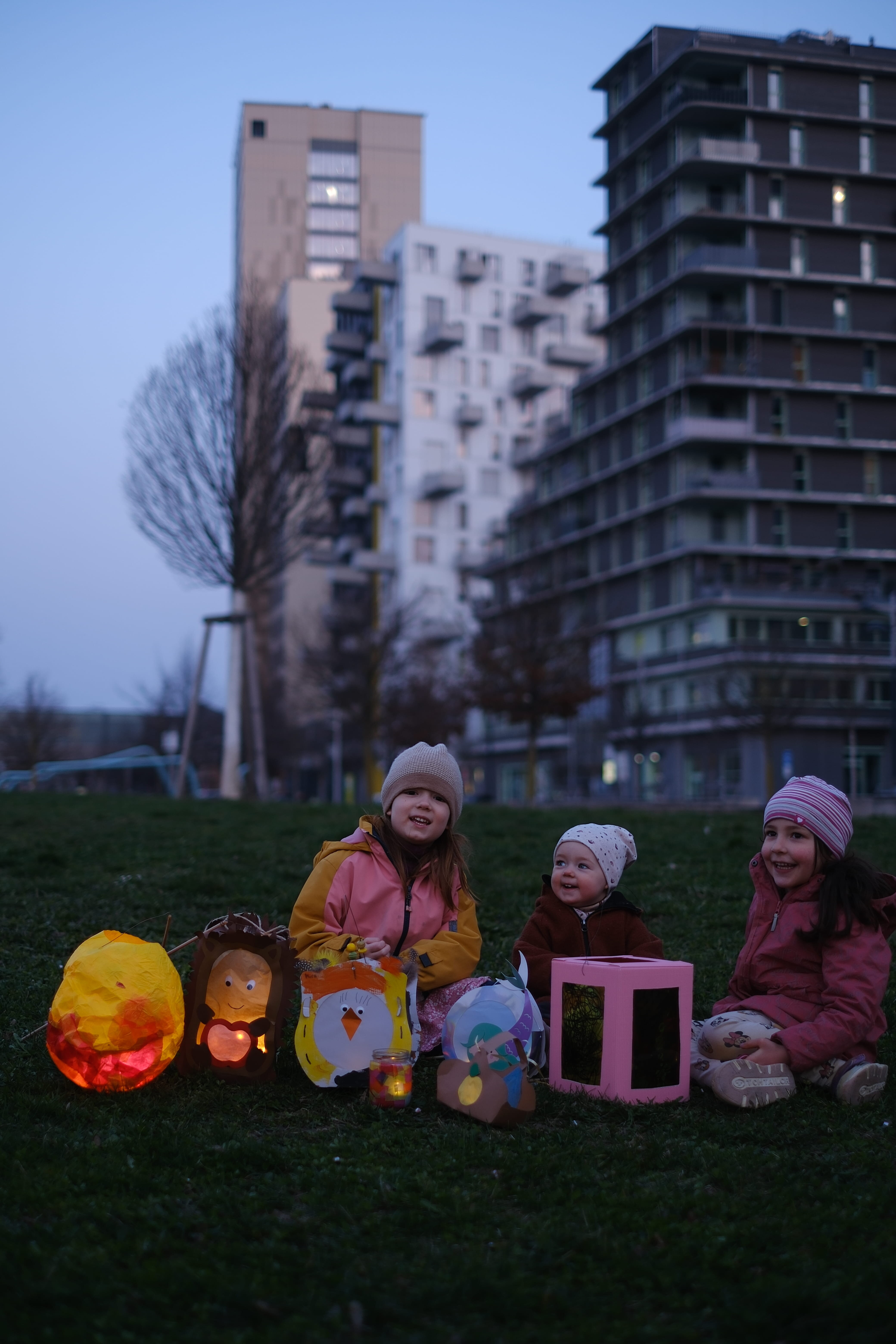 Drei Kinder sitzen im Gras, es stehen leuchtende Laternen vor ihnen