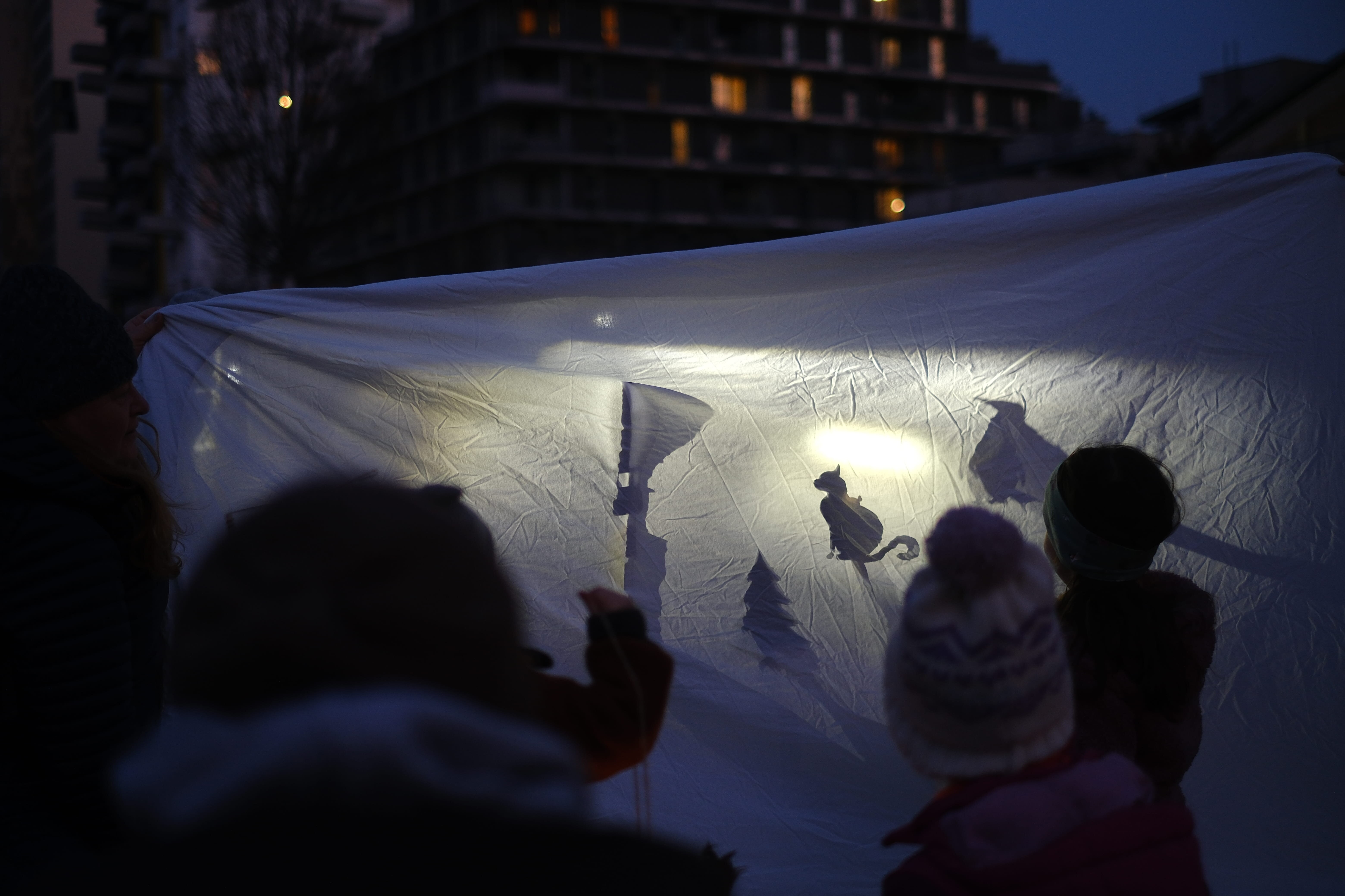 Schattentheater im Freien mit einem weißen Leintuch und Taschenlampen, zwei Kinder sitzen davor und schauen zu