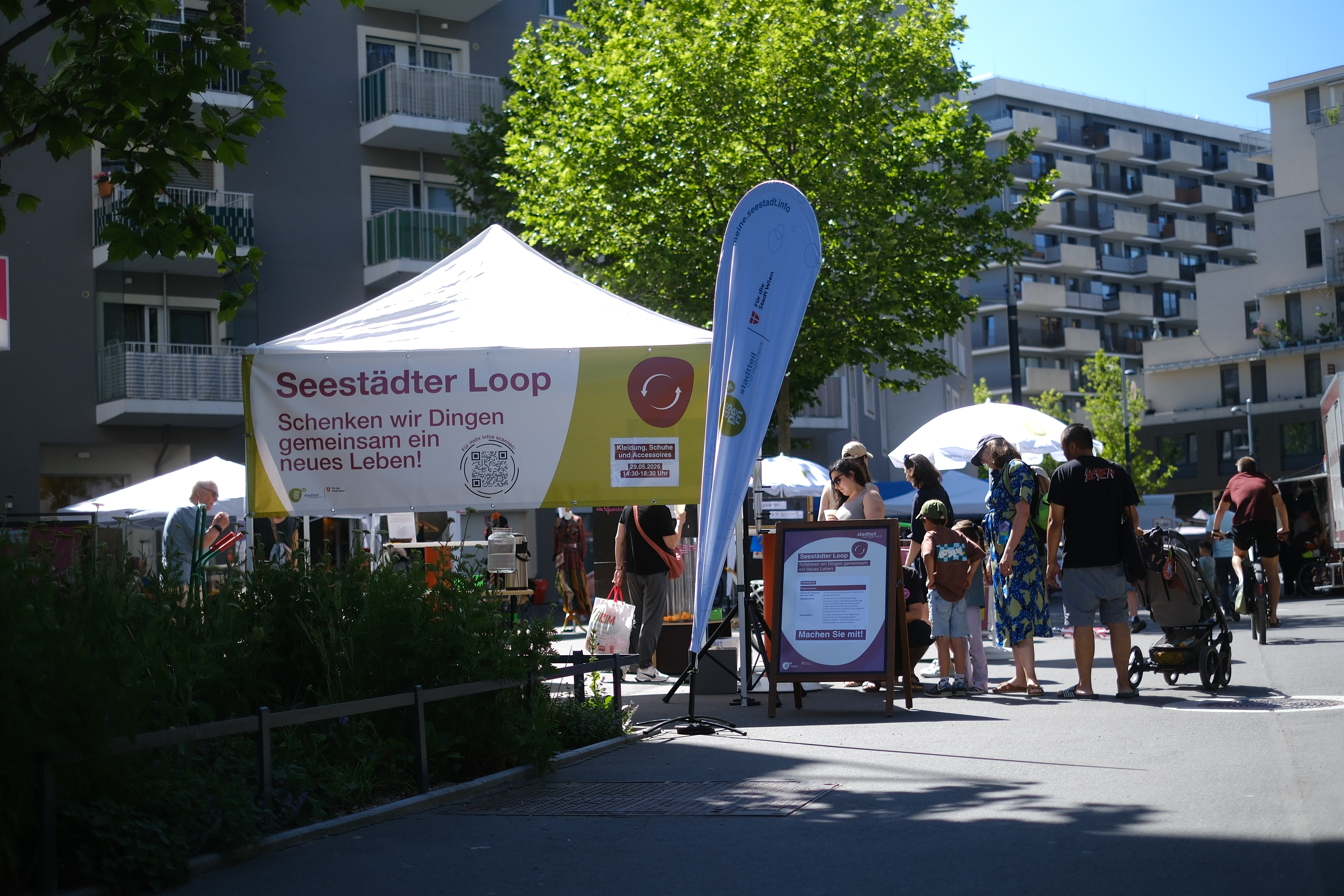 Platz mit Menschen, die bei einem Pavillon stehen und auf dem ein großer Banner mit der Aufschrift "Seestädter Loop. Schenken wir Dingen gemeinsam ein neues Leben" steht.