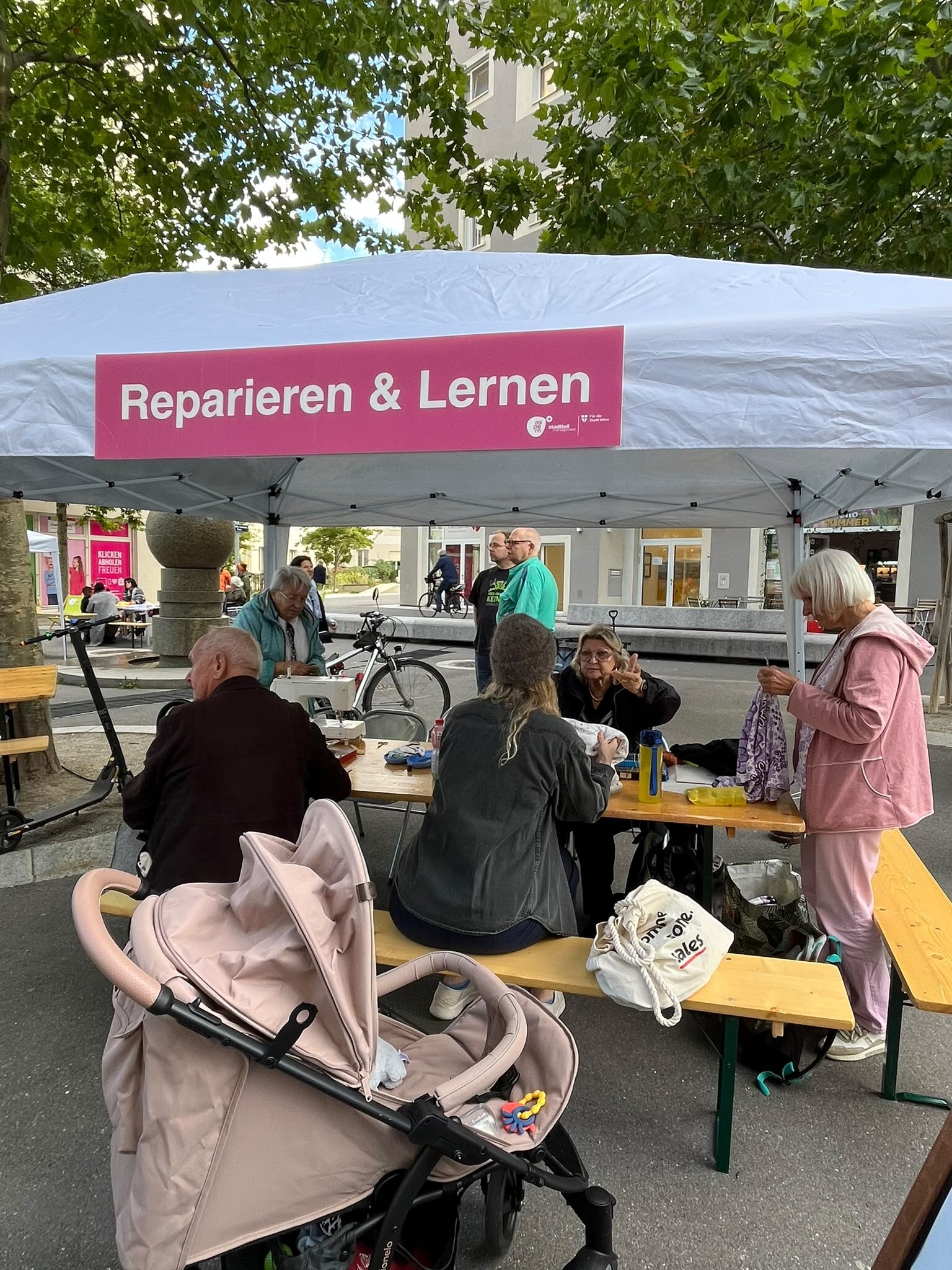 Personen sitzen auf Bänken unter einem Pavillon mit der Aufschrift Reparieren & Lernen